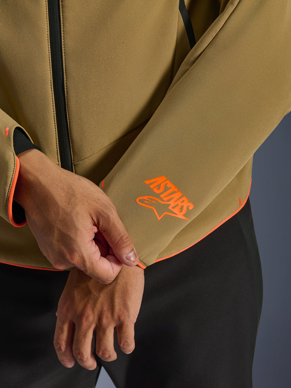 Alpinestars Aeroshell Waterproof Jacket, kolor ciemny khaki z pomarańczowymi akcentami fluo, zbliżenie na mankiet rękawa i dolną krawędź z jaskrawopomarańczowym logo Astars, neonową lamówką i wodoodpornym zamkiem błyskawicznym, sportowa konstrukcja softshell.