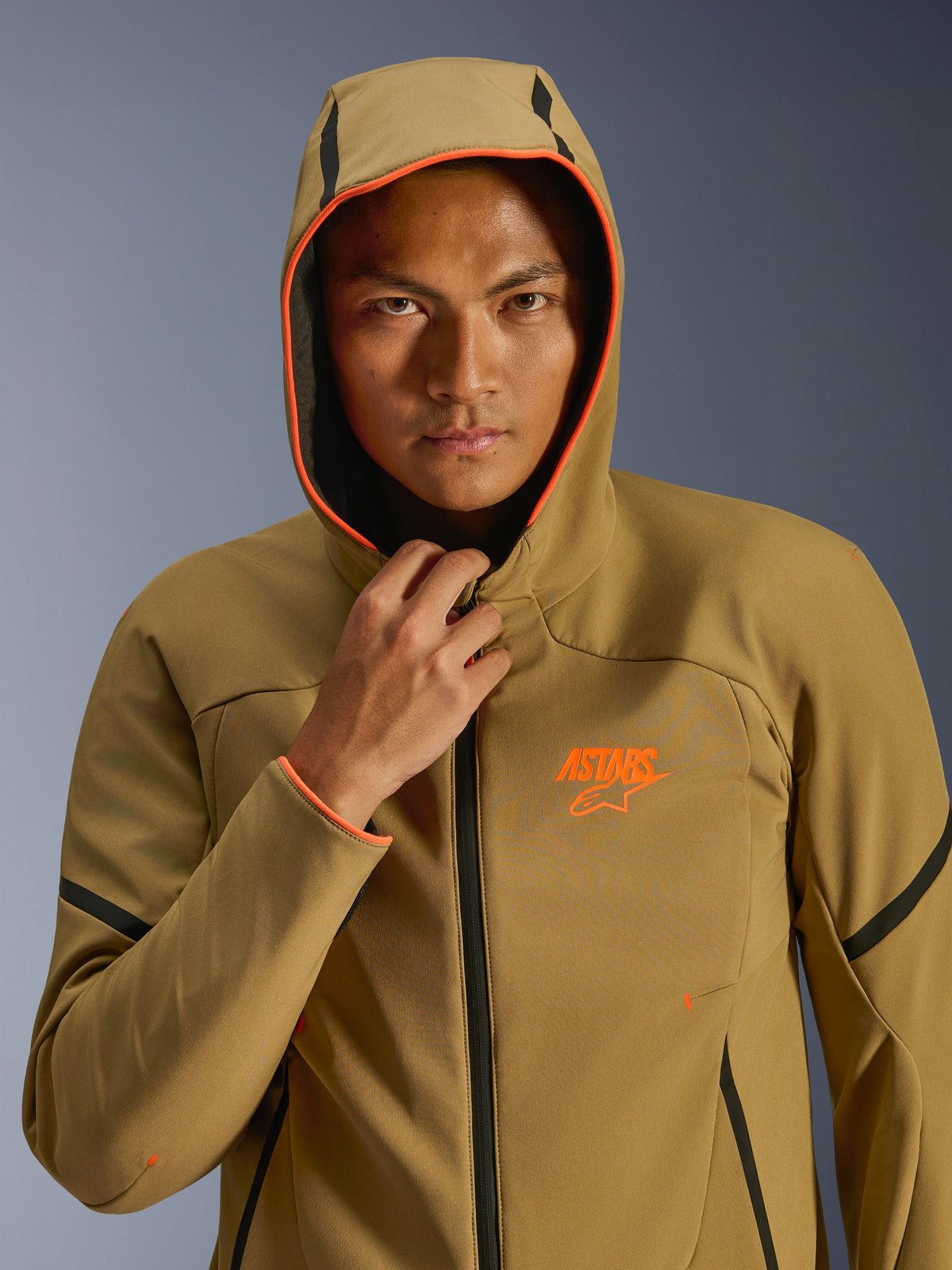 Kurtka Alpinestars Aeroshell Waterproof, Dark Khaki Black Orange Fluo, model mający na sobie kurtkę z założonym kapturem, jaskrawe neonowe pomarańczowe wykończenie i pomarańczowe logo ASTARS, czarne akcenty, nowoczesny wodoodporny softshell w sportowym stylu