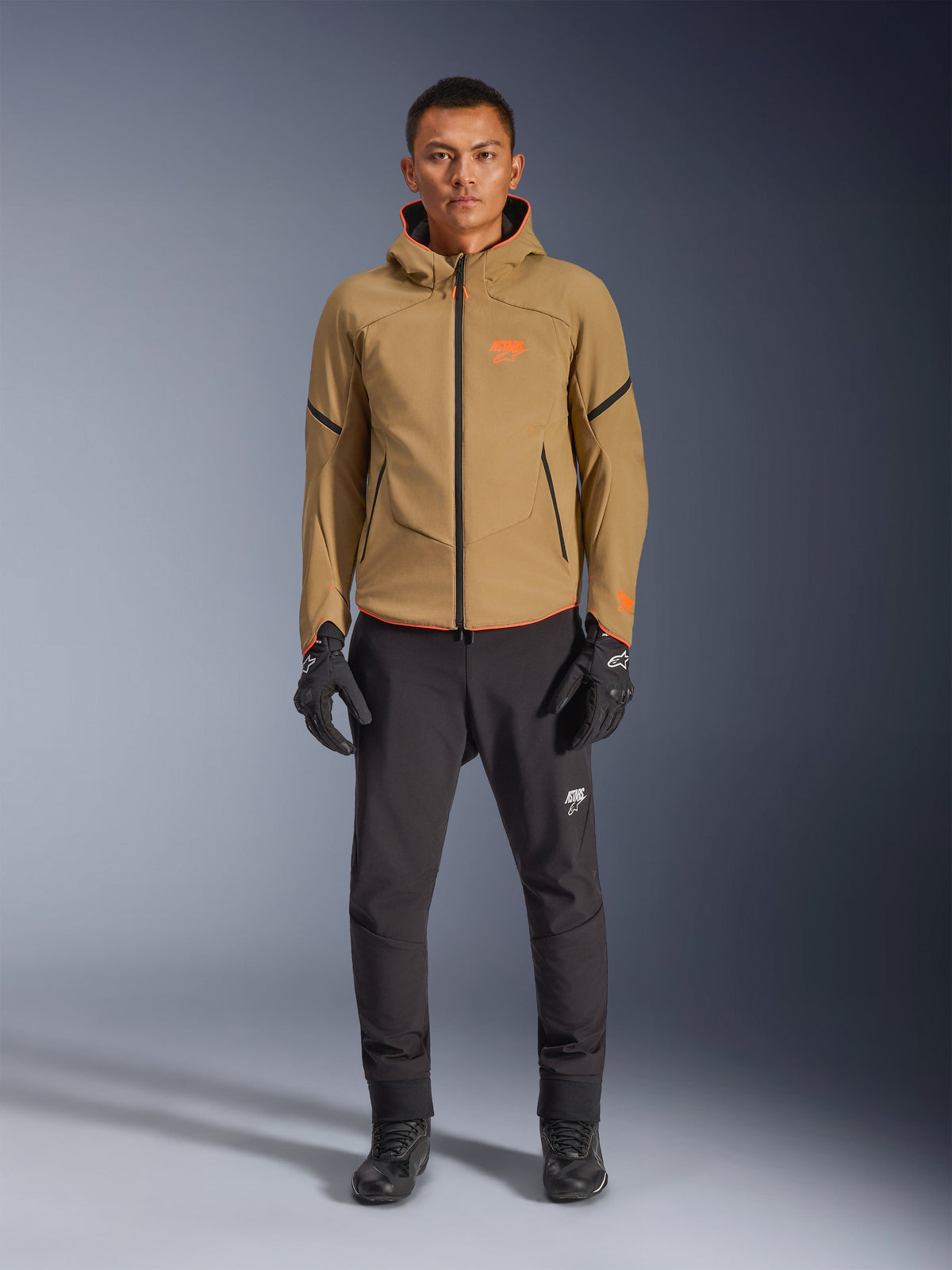 Męski model w kurtce Alpinestars Aeroshell Waterproof w kolorze Dark Khaki z czarnymi i pomarańczowymi akcentami fluo, pomarańczowe logo na piersi, design z kapturem, czarne spodnie i czarne rękawice motocyklowe, styl casual sport-touring