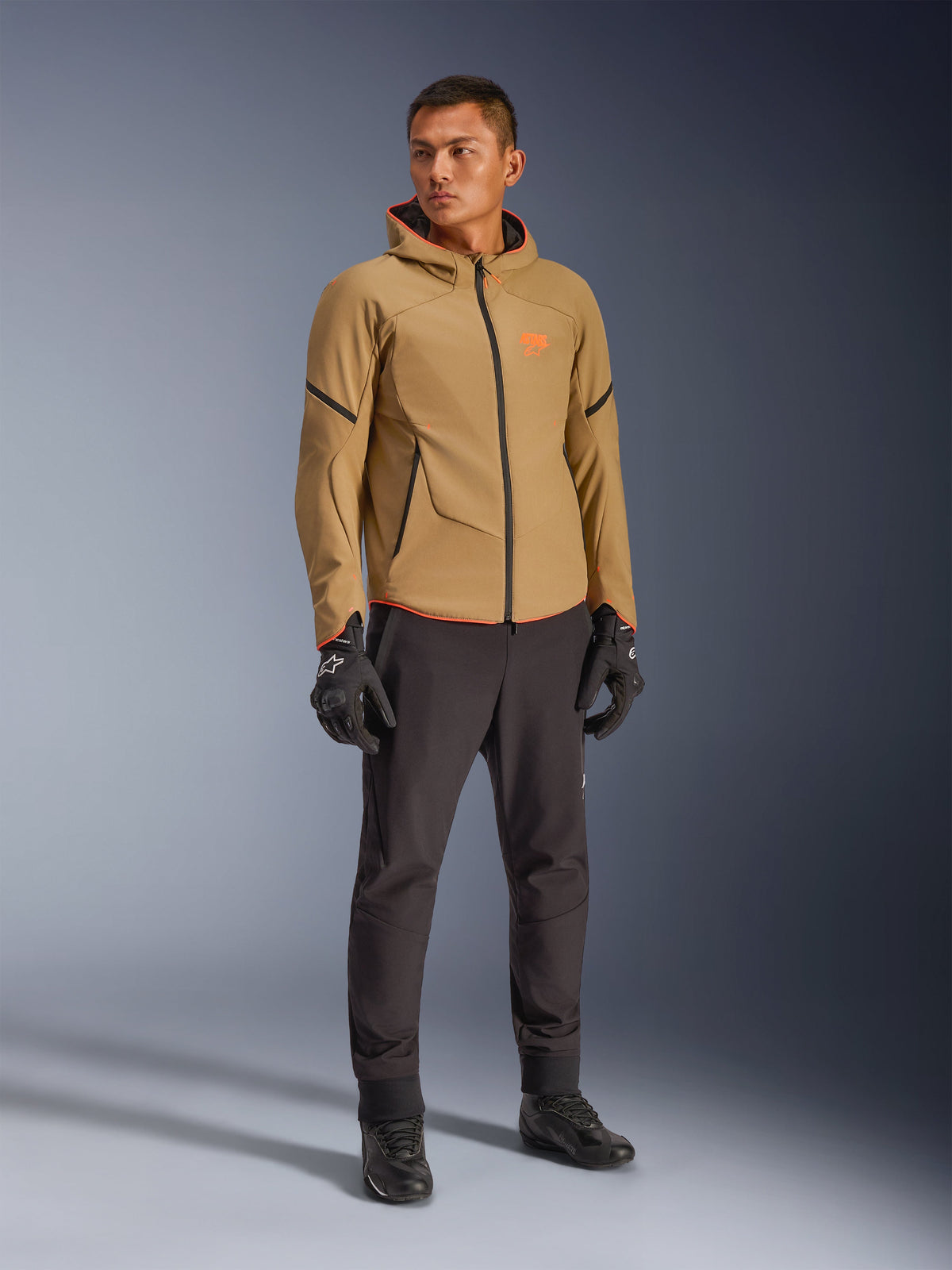 Wodoodporna kurtka Alpinestars Aeroshell w kolorach Dark Khaki, czarnym i jaskrawym pomarańczowym (Orange Fluo), noszona przez modela z dopasowaną odzieżą, konstrukcja softshell z kapturem, wodoodporne zamki, jaskrawopomarańczowe akcenty i branding.