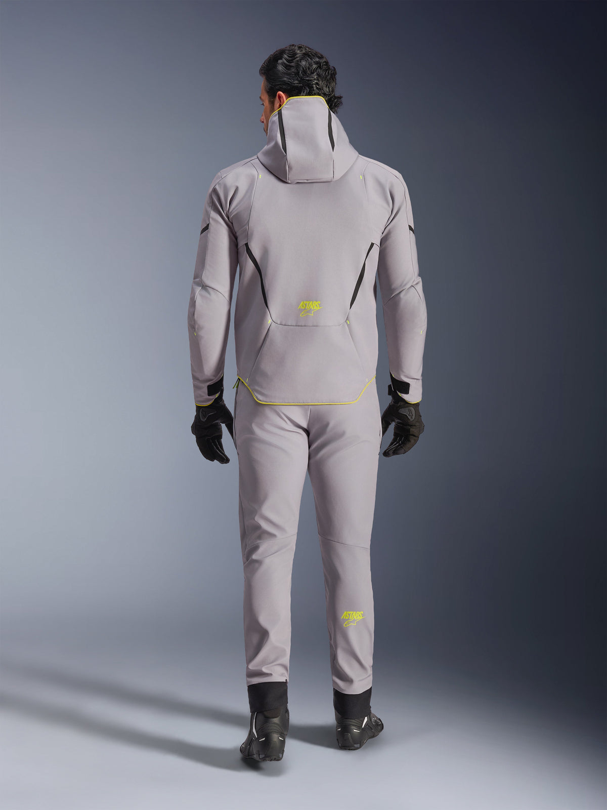 Wodoodporna kurtka Alpinestars Aeroshell w kolorze Mid Gray Yellow Fluo, pełny widok z tyłu, noszona z pasującymi szarymi spodniami, czarnymi rękawicami i czarnymi butami motocyklowymi. Posiada konstrukcję softshell z kapturem, jaskrawożółte akcenty oraz logo 'ASTARS', zaprojektowana z myślą o nowoczesnych osiągach motocyklowych na drodze.