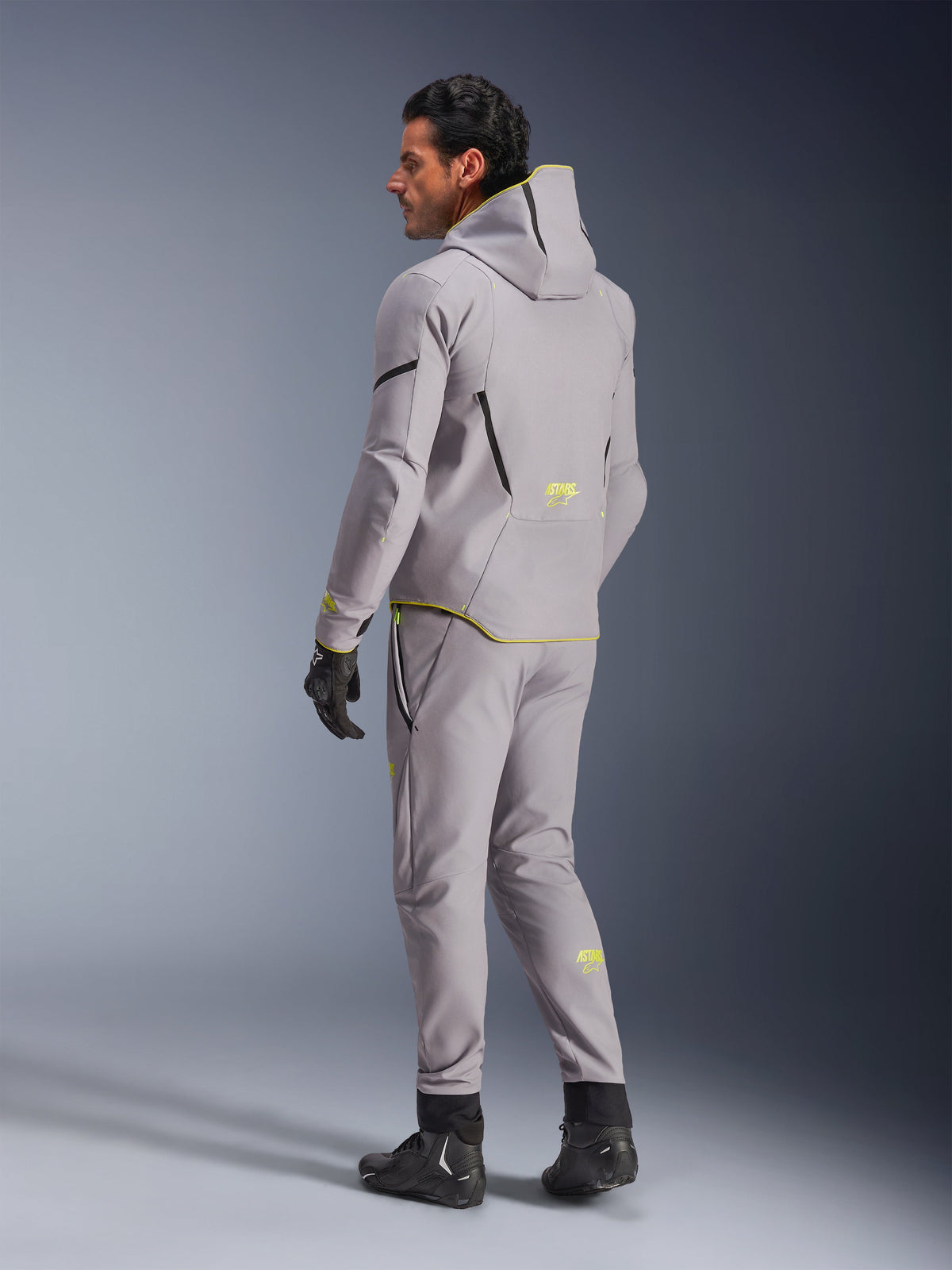 Wodoodporna kurtka Alpinestars Aeroshell, kolor Mid Gray Yellow Fluo, widok z tyłu, design bluzy z kapturem z żółtymi akcentami i logo, zestawiona z pasującymi szarymi spodniami, czarnymi rękawicami i butami motocyklowymi, nowoczesna odzież motocyklowa