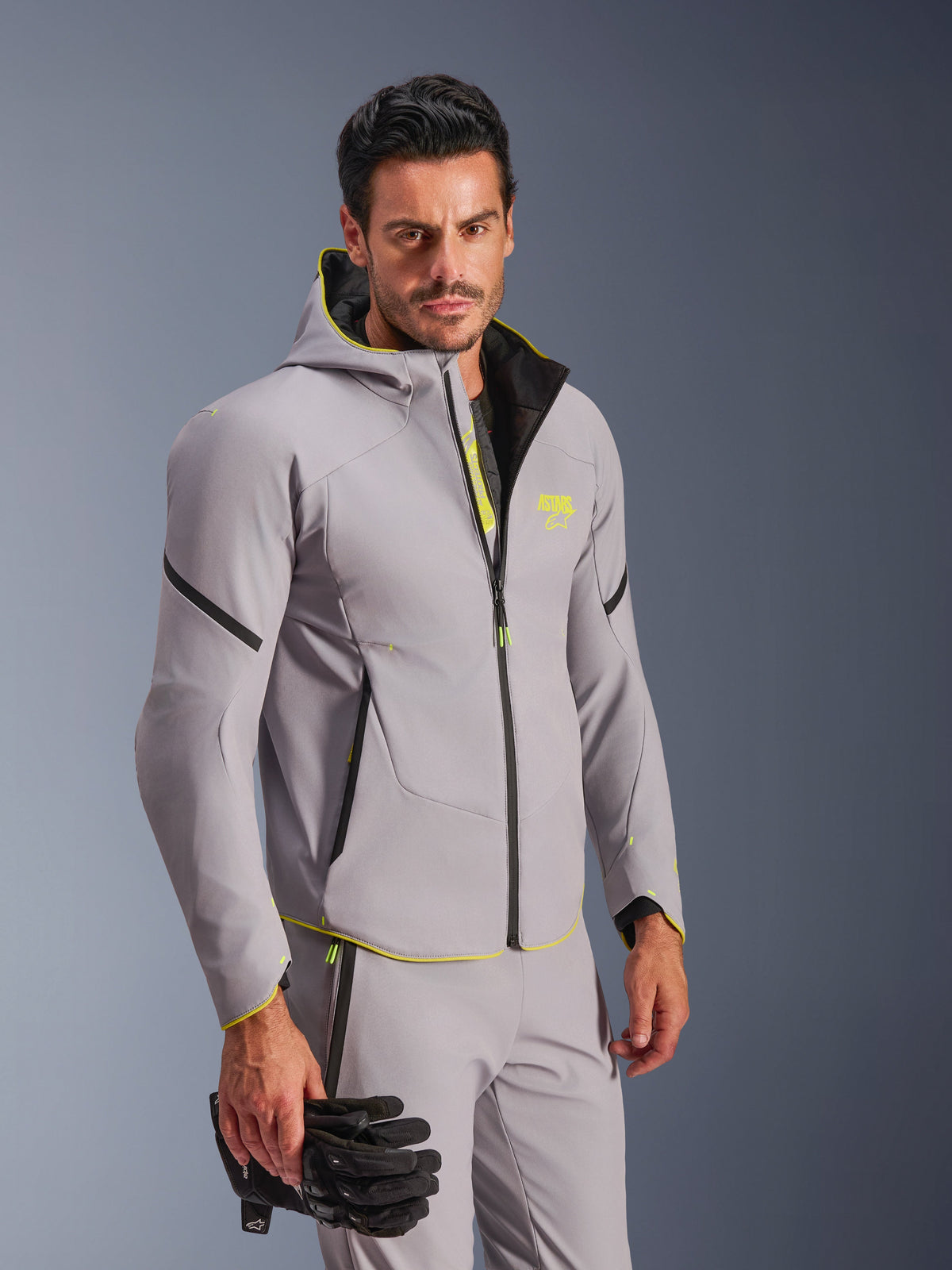 Alpinestars Aeroshell Waterproof Jacket w kolorze szarym i neonowym żółtym, noszona przez mężczyznę z pasującymi szarymi spodniami i czarnymi rękawicami motocyklowymi, fason softshell z kapturem z neonowożółtymi akcentami i logo na piersi, nowoczesna sportowa odzież motocyklowa.