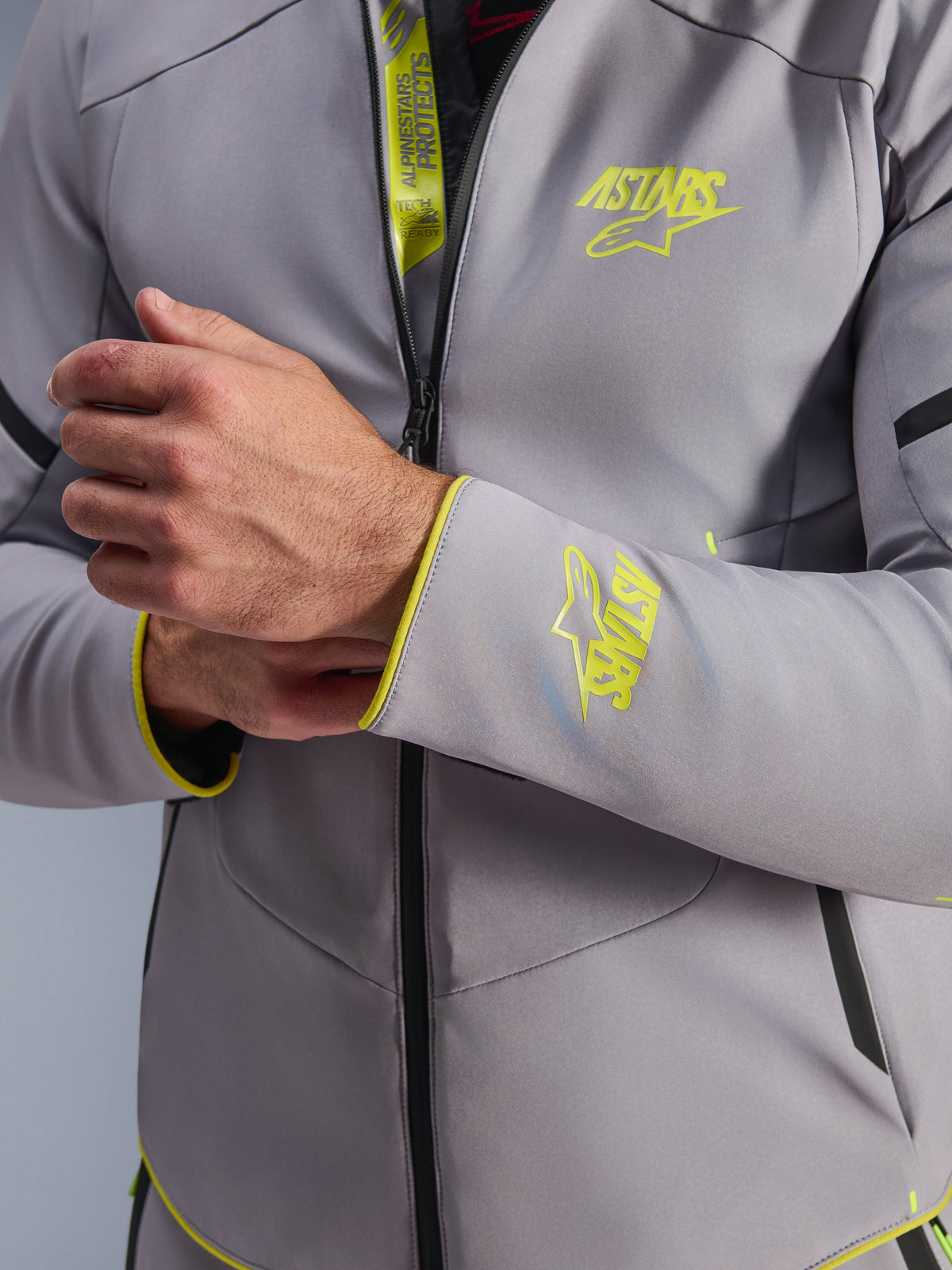 Alpinestars Aeroshell Waterproof Jacket, szara z jaskrawożółtymi akcentami fluo, zbliżenie na klatkę piersiową i mankiet rękawa z jaskrawożółtym logo Astars, detal wewnętrznej klapy z oznaczeniem Tech-Air Ready, nowoczesna sportowa motocyklowa kurtka softshell
