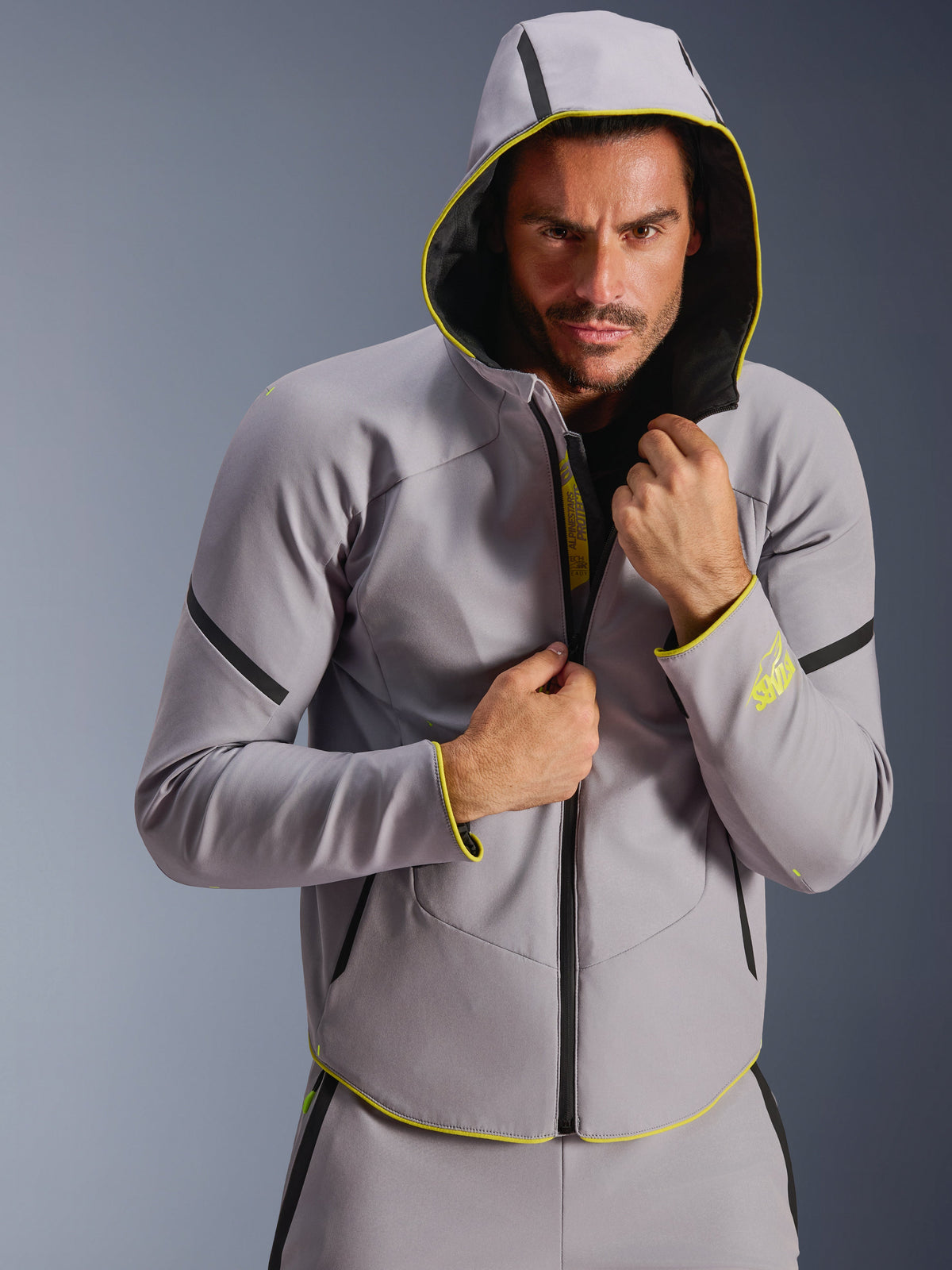 Alpinestars Aeroshell Waterproof Jacket, szaro-żółta (Mid Gray i Yellow Fluo), sportowa kurtka softshell z kapturem, noszona przez modela z założonym kapturem, czarne wodoodporne zamki i neonowożółte akcenty na mankietach oraz dolnej krawędzi, nowoczesny design inspirowany motorsportem