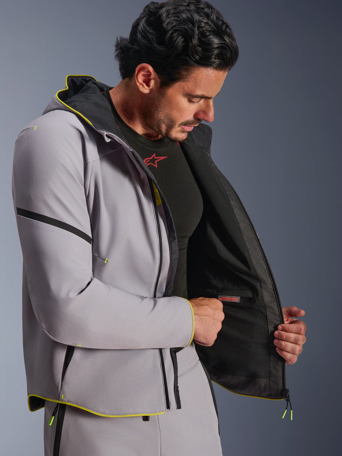 Kurtka Alpinestars Aeroshell Waterproof w kolorze szarym i neonowym żółtym, model prezentujący wewnętrzną wodoodporną komorę oraz czarną podszewkę, sportowa motocyklowa bluza softshell z kapturem, neonowe żółte wykończenia, nowoczesny sportowy krój