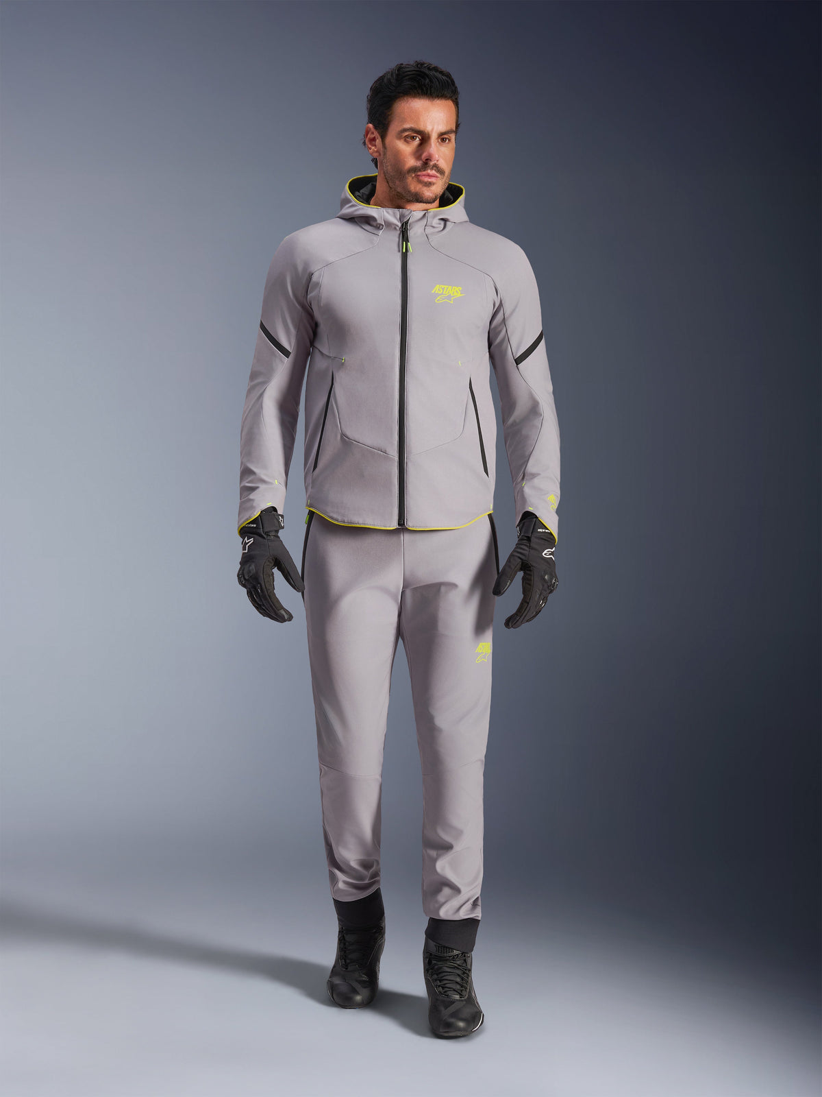 Alpinestars Aeroshell Waterproof Jacket, Mid Gray Yellow Fluo, noszona z pasującymi szarymi spodniami, czarnymi rękawicami i butami, o nowoczesnym sportowym designie bluzy z kapturem z jaskrawożółtymi akcentami i sportowym dopasowaniu.