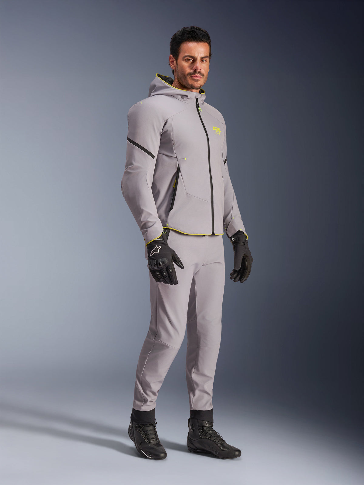 Kurtka Alpinestars Aeroshell Waterproof, szara i neonowa żółta, prezentowana przez modela z pasującymi szarymi spodniami, czarnymi rękawicami motocyklowymi i czarnymi butami, nowoczesna odzież motocyklowa w stylu bluzy z kapturem do jazdy miejskiej i sportowej.