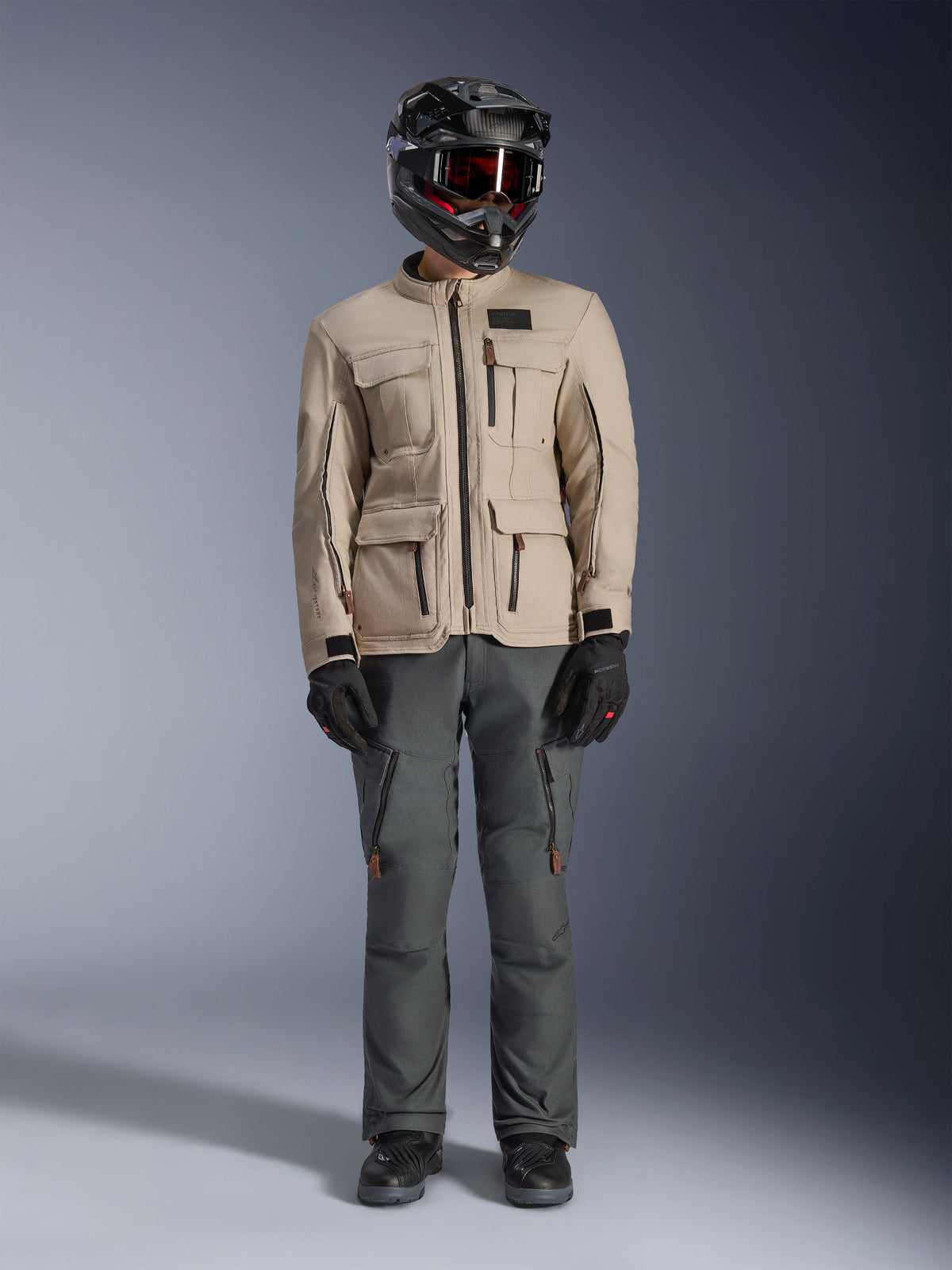 Alpinestars Borrego Drystar® Jacket in Warm Gray, adventure motorcycle jacket with four front cargo pockets, paired with dark gray riding pants, black off-road helmet, and black gloves, featuring a rugged waxed-canvas look