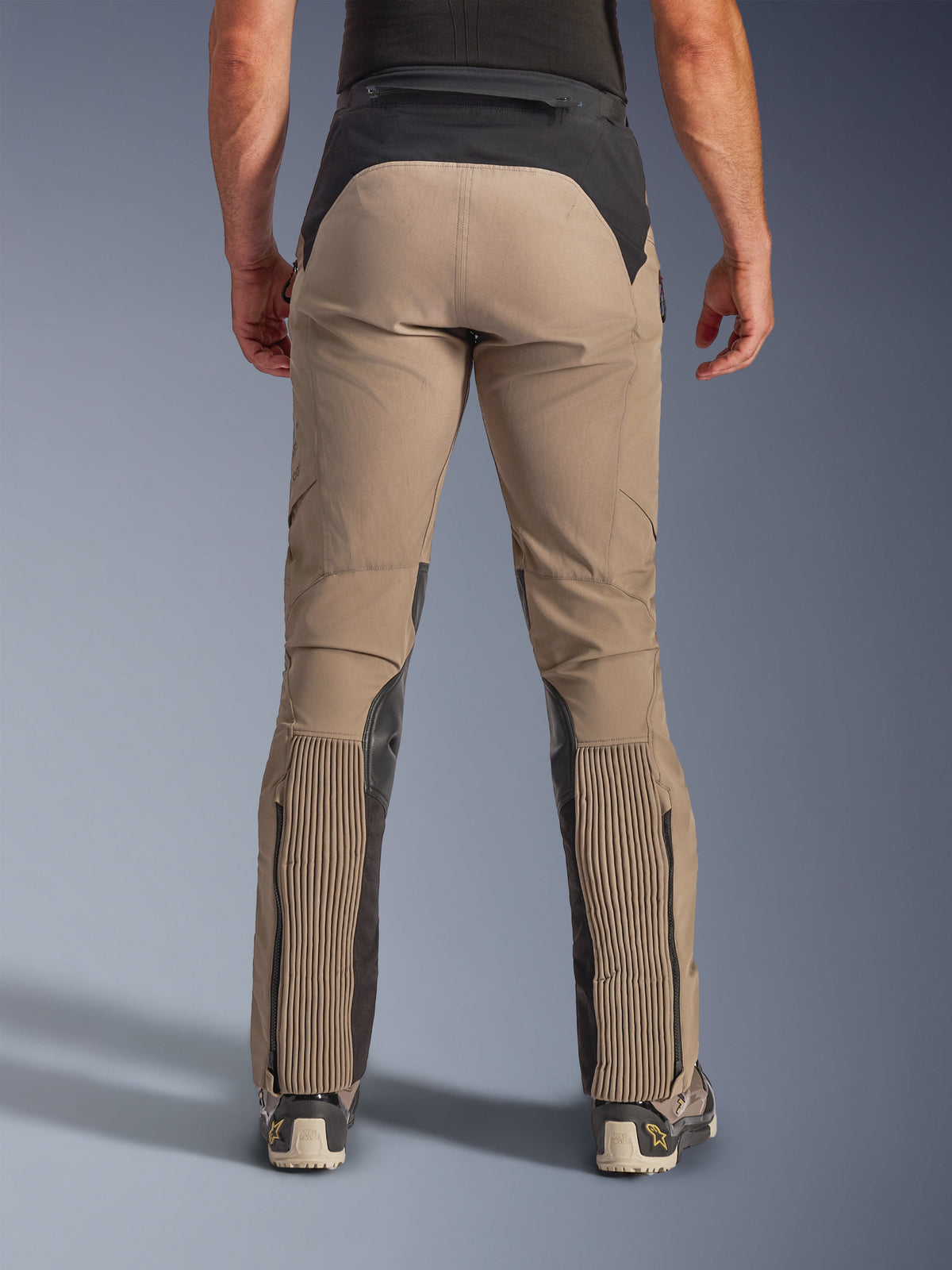 Spodnie Alpinestars AMT-8 Stretch Drystar® XF, Walnut Black, widok z tyłu na modelu, beżowa elastyczna tkanina z czarnym karczkiem i panelami na łydkach, duże akordeonowe sekcje elastyczne w dolnej części nogawek, nogawki zapinane na zamek, krój adventure-touring