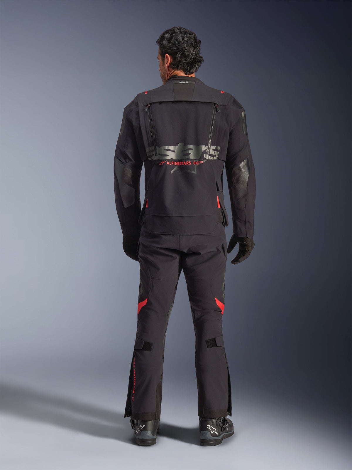 Motocyklista widoczny od tyłu ubrany w kurtkę Alpinestars Halo Pro Drystar® XF Laminated w kolorze Black Dark Shadow, z dużym szarym logo astars i czerwonym brandingiem Alpinestars, modułowymi panelami wentylacyjnymi na plecach, w zestawie z pasującymi spodniami przygodowymi i czarnymi butami.