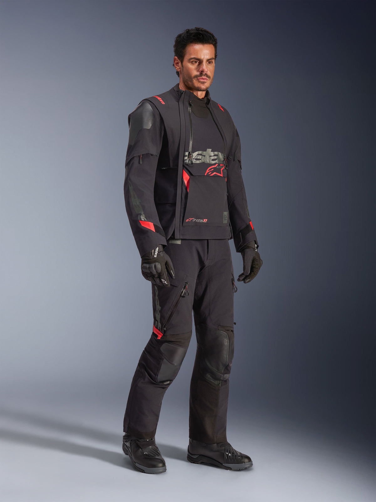 Motocyklista w kurtce Alpinestars Halo Pro Drystar® XF Laminated Jacket w kolorze Black Dark Shadow, kompletny strój adventure z dopasowanymi spodniami, rękawicami i butami, modułowe asymetryczne panele przednie i czerwone akcenty brandingowe