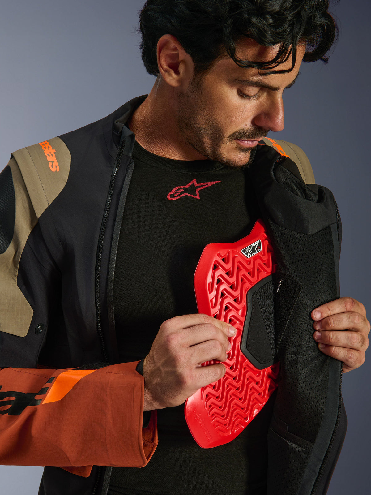 Kurtka Alpinestars Halo Pro Drystar® XF Laminated w kolorze Walnut Rust Black, zdjęcie lifestylowe modela rozpinającego kurtkę adventure, aby odsłonić czerwoną wkładkę ochraniacza klatki piersiowej, pomarańczowy branding na ramieniu i czarna warstwa bazowa z czerwonym logo.
