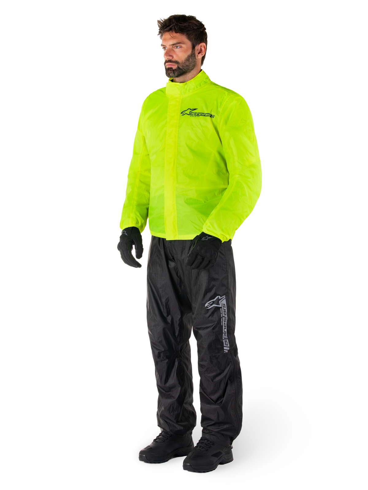 Mężczyzna ubrany w kurtkę Alpinestars Hurricane Rain V2 w kolorze Yellow Fluo i spodnie Hurricane Rain V2 w kolorze czarnym, wodoodporna motocyklowa odzież przeciwdeszczowa o wysokiej widoczności, odblaskowe logo na klatce piersiowej i udzie, kompletny zestaw z czarnymi rękawicami i butami.
