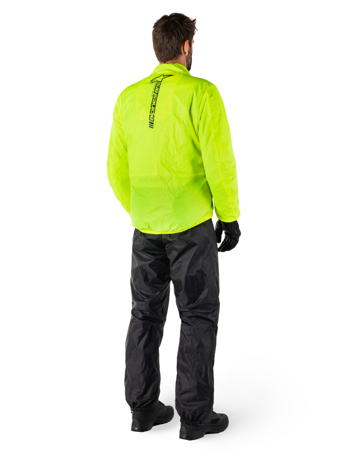 Osoba widoczna od tyłu ubrana w kurtkę Alpinestars Hurricane Rain V2 w kolorze Yellow Fluo i spodnie Hurricane Rain V2 w kolorze czarnym, pionowe logo na plecach kurtki, wodoodporna motocyklowa odzież przeciwdeszczowa, czarne rękawice i buty