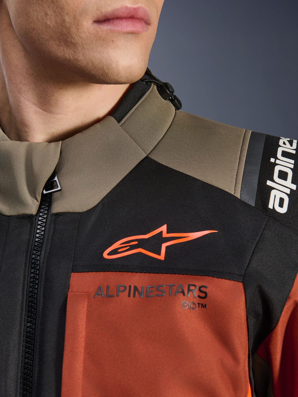 Kurtka Alpinestars Andes V4 Drystar® w kolorze Walnut Rust Black, zbliżenie na górną część klatki piersiowej i ramię, pomarańczowe logo A-star na czarnym panelu i napis Alpinestars na rdzawym elemencie, detal motocyklowej odzieży typu adventure