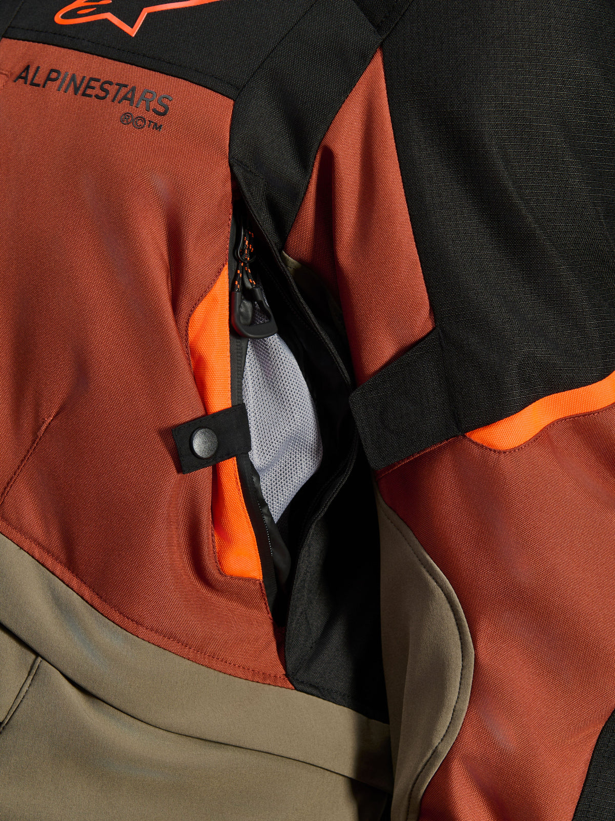 Alpinestars Andes V4 Drystar® Jacket, Walnut Rust Black, zbliżenie na bezpośredni otwór wentylacyjny z siatkową podszewką, wytrzymały materiał tekstylny z pomarańczowymi akcentami, zatrzask i detal z logo Alpinestars