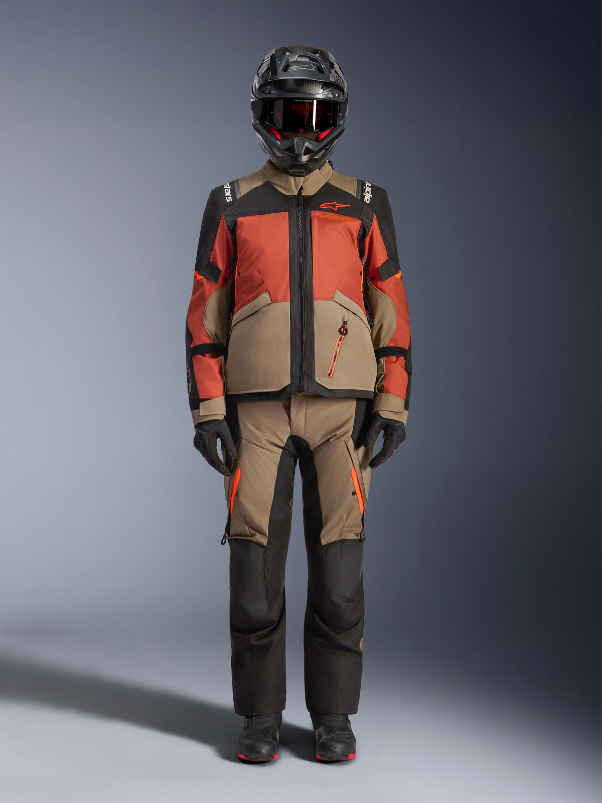 Motocyklista przygodowy ubrany w kurtkę Alpinestars Andes V4 Drystar® w kolorze Walnut Rust Black, zestawioną z pasującymi spodniami adventure, czarnym kaskiem z czerwonymi akcentami i goglami oraz czarnymi rękawicami, prezentujący kompletny strój turystyczny na każdą pogodę.