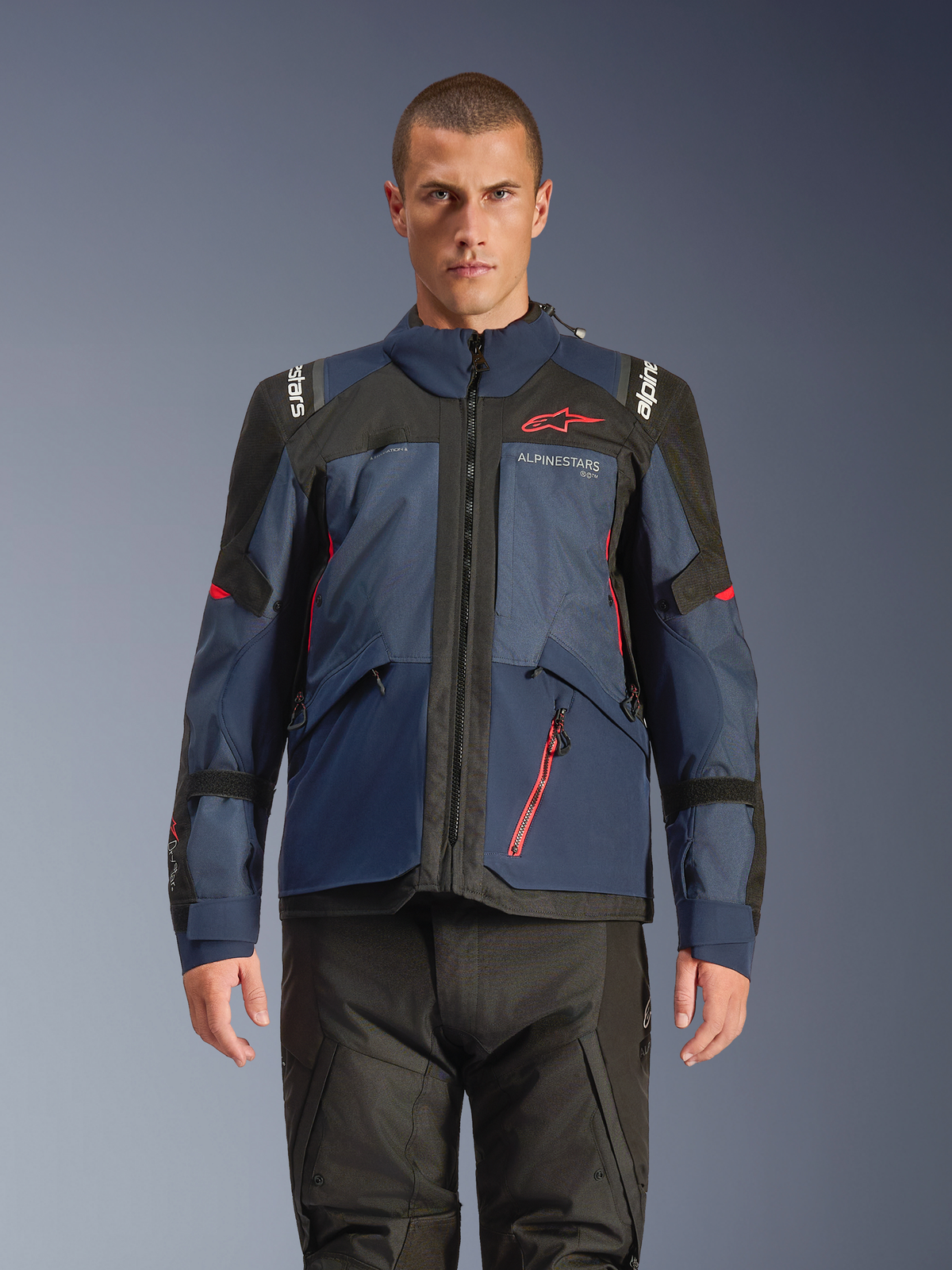 Kurtka Alpinestars Andes V4 Drystar®, przygodowa kurtka motocyklowa, kolory Night Navy i Mood Indigo z czarnymi panelami, prezentowana przez modela w czarnych spodniach motocyklowych, czerwone logo Alpinestars na piersi i czerwone detale zamka, wytrzymała konstrukcja tekstylna do turystyki w każdych warunkach pogodowych
