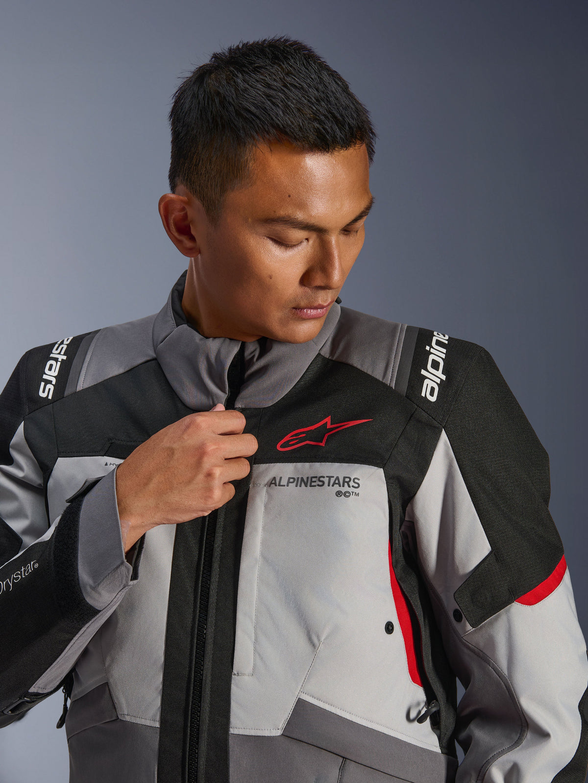 Model wearing Alpinestars Andes V4 Drystar® Jacket, Dark Gray and Ice Gray with Black and Bright Red accents, adjusting the front zipper, featuring the red Astars logo and Drystar® branding, adventure touring style
