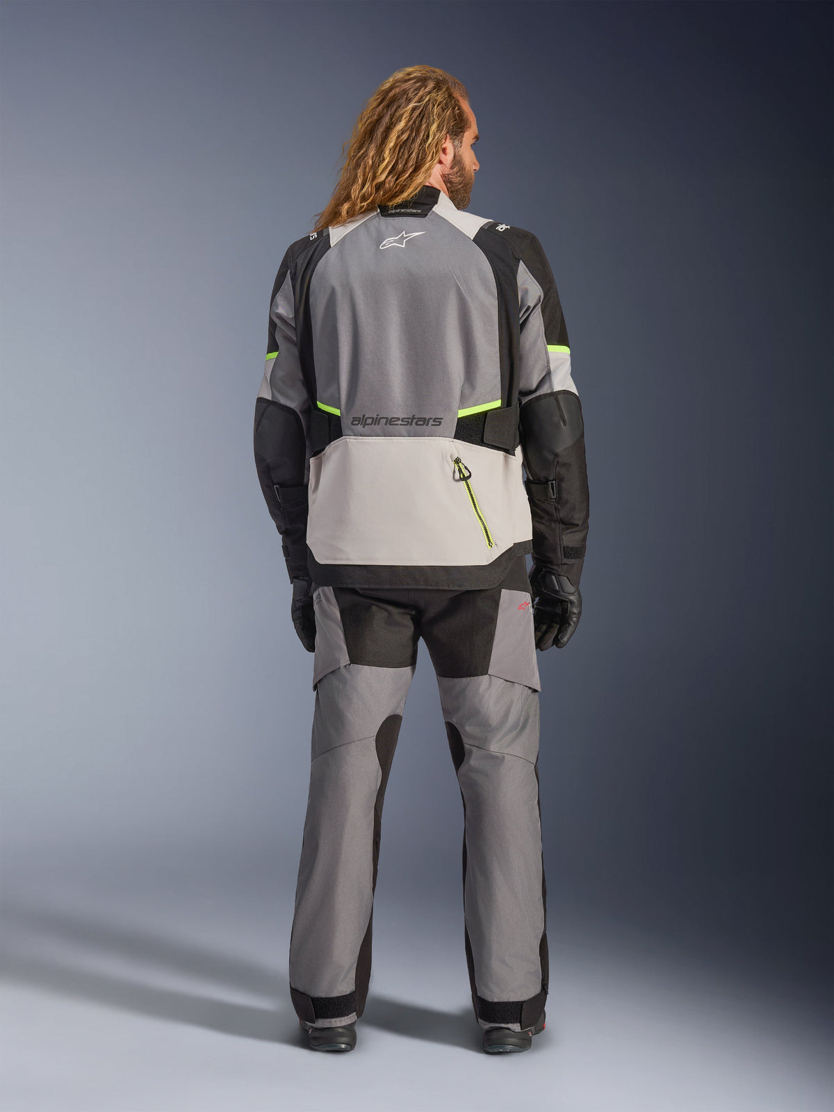Kurtka Alpinestars Andes V4 Drystar®, motocyklowa kurtka adventure, ciemnoszara i jasnoszara z czarnymi i neonowożółtymi akcentami, widok z tyłu w zestawie z pasującymi spodniami i czarnymi rękawicami, duża tylna kieszeń z żółtym zamkiem i wzmocnione panele na ramionach i łokciach
