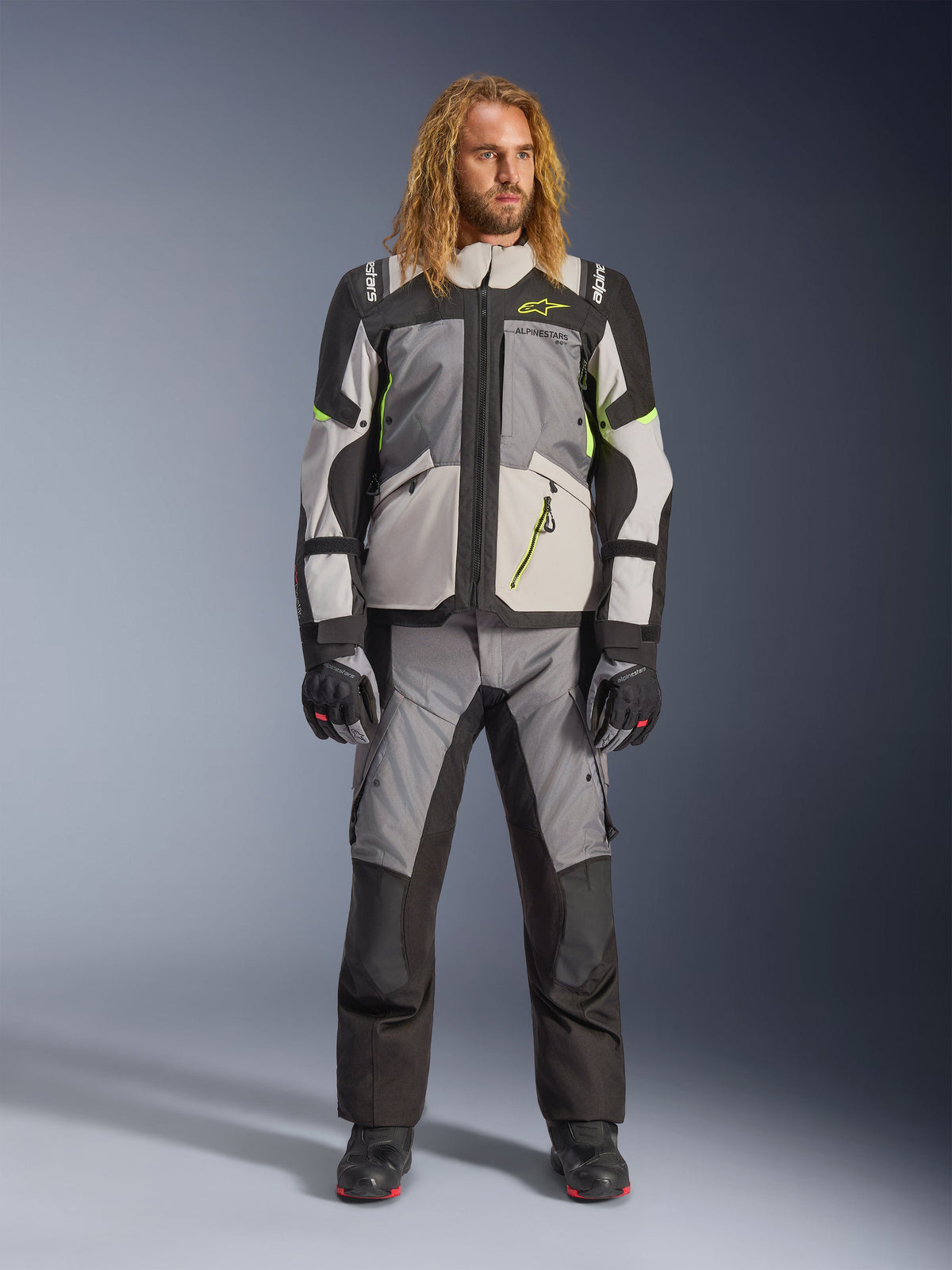 Męski model z długimi blond włosami ubrany w kurtkę Andes V4 Drystar® w kolorach ciemnoszarym, jasnoszarym, czarnym i żółtym fluo, z dopasowanymi spodniami motocyklowymi adventure i czarnymi butami, kompletny techniczny strój motocyklowy do turystyki w każdych warunkach pogodowych.