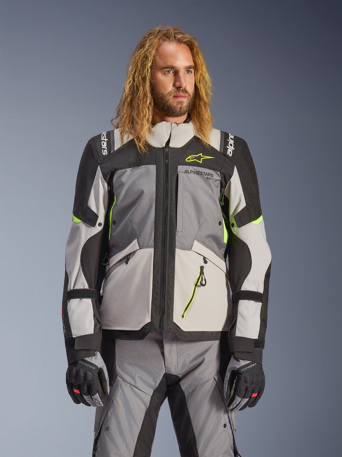 Alpinestars Andes V4 Drystar Jacket, turystyczna kurtka motocyklowa adventure, ciemnoszara i jasnoszara z neonowożółtymi akcentami, noszona przez modela z pasującymi spodniami i rękawicami, tekstylna konstrukcja z wieloma kieszeniami do jazdy w każdych warunkach pogodowych