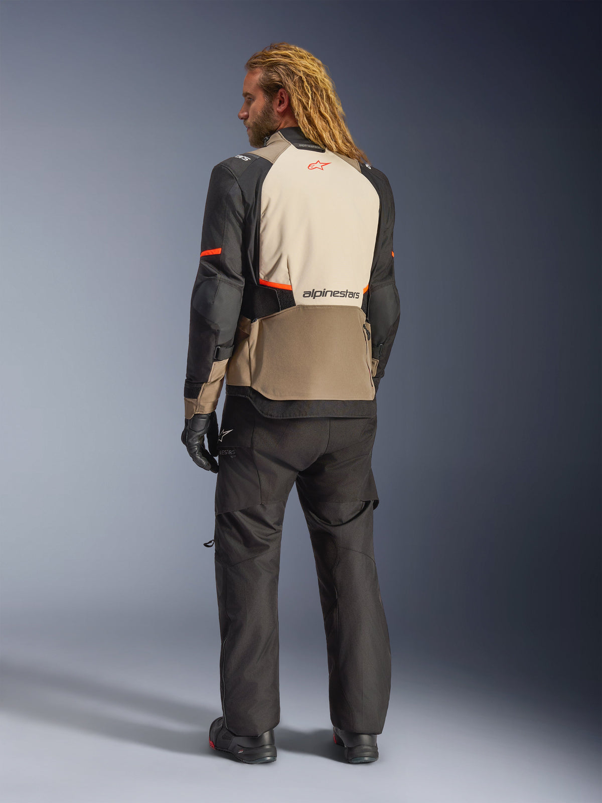 Kurtka Alpinestars Andes V4 Drystar®, Walnut Mastic Black, widok z tyłu na modelu, beżowe i czarne wzmocnione panele tekstylne z pomarańczowymi akcentami, logo Alpinestars na plecach, noszona z dopasowanymi czarnymi spodniami motocyklowymi adventure i rękawicami.