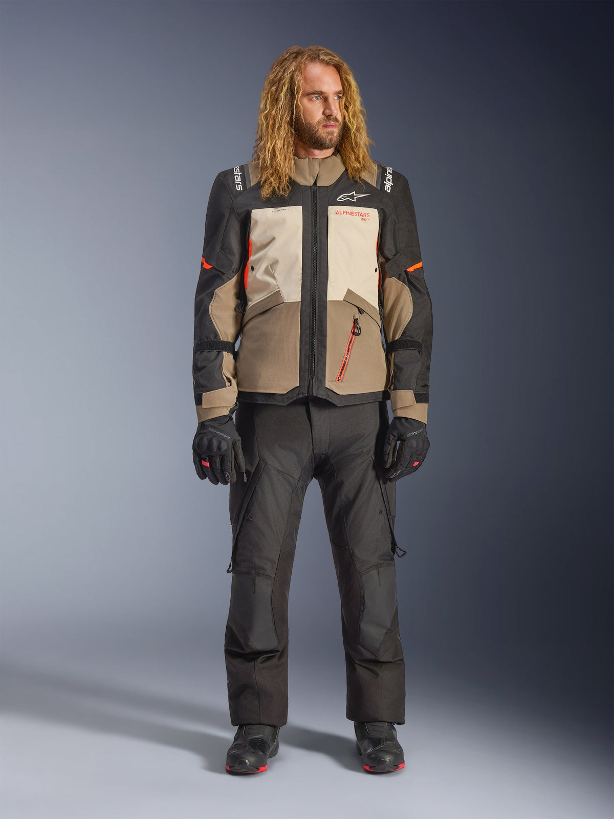 Kurtka Alpinestars Andes V4 Drystar®, Walnut Mastic Black, noszona z czarnymi spodniami motocyklowymi i rękawicami, wielopanelowa konstrukcja adventure z pomarańczowymi akcentami i brandingiem Alpinestars, techniczna odzież tekstylna do jazdy w każdych warunkach pogodowych
