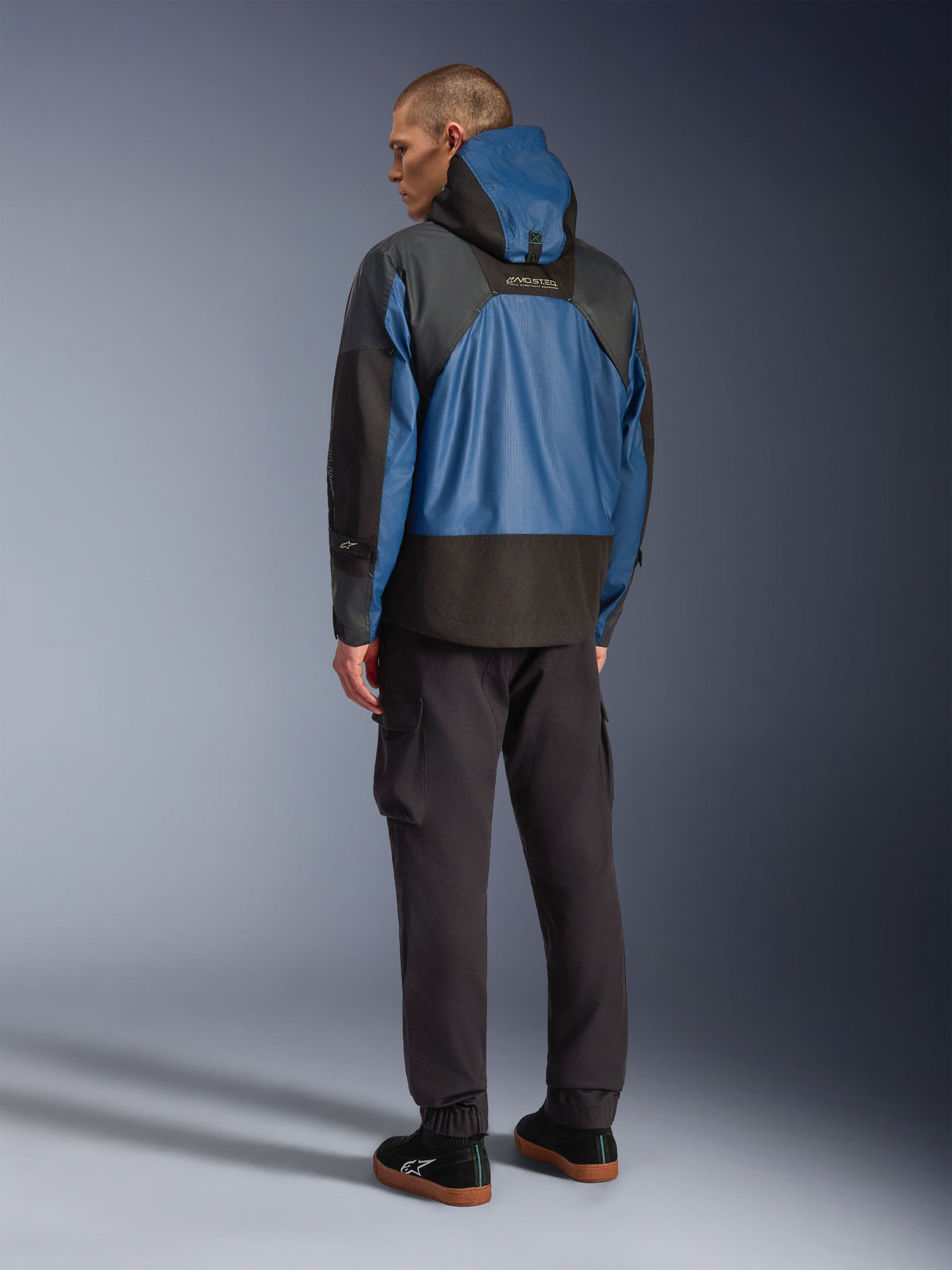 Rear view of a male model wearing the Alpinestars Mospher Waterproof Jacket in Black and Dark Petrol, featuring a hooded design and tactical ripstop construction, paired with black cargo pants and sneakers.