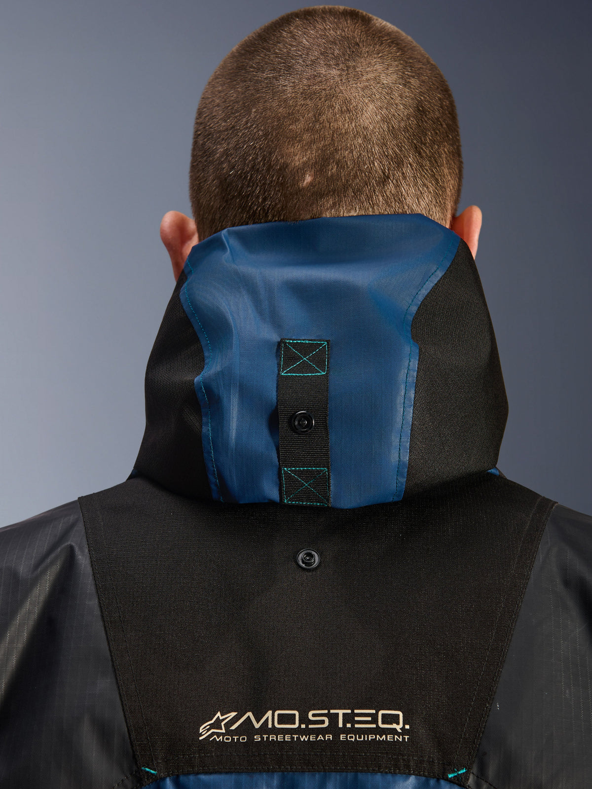 Alpinestars Mospher Waterproof Jacket, back view detail showing the hood anchoring system, Black and Dark Petrol Blue, featuring MO.ST.EQ. Moto Streetwear Equipment branding, durable ripstop textile material, technical commuter motorcycle gear
