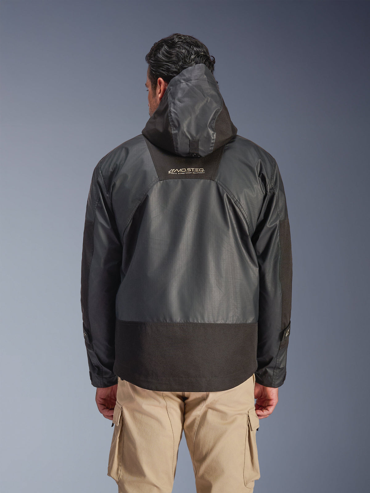 Widok z tyłu mężczyzny w czarnej kurtce Alpinestars Mospher Waterproof Jacket z kapturem, trwała konstrukcja z tkaniny ripstop, beżowe spodnie cargo, widoczne logo MO.ST.EQ. na górze pleców, miejski styl motocyklowy