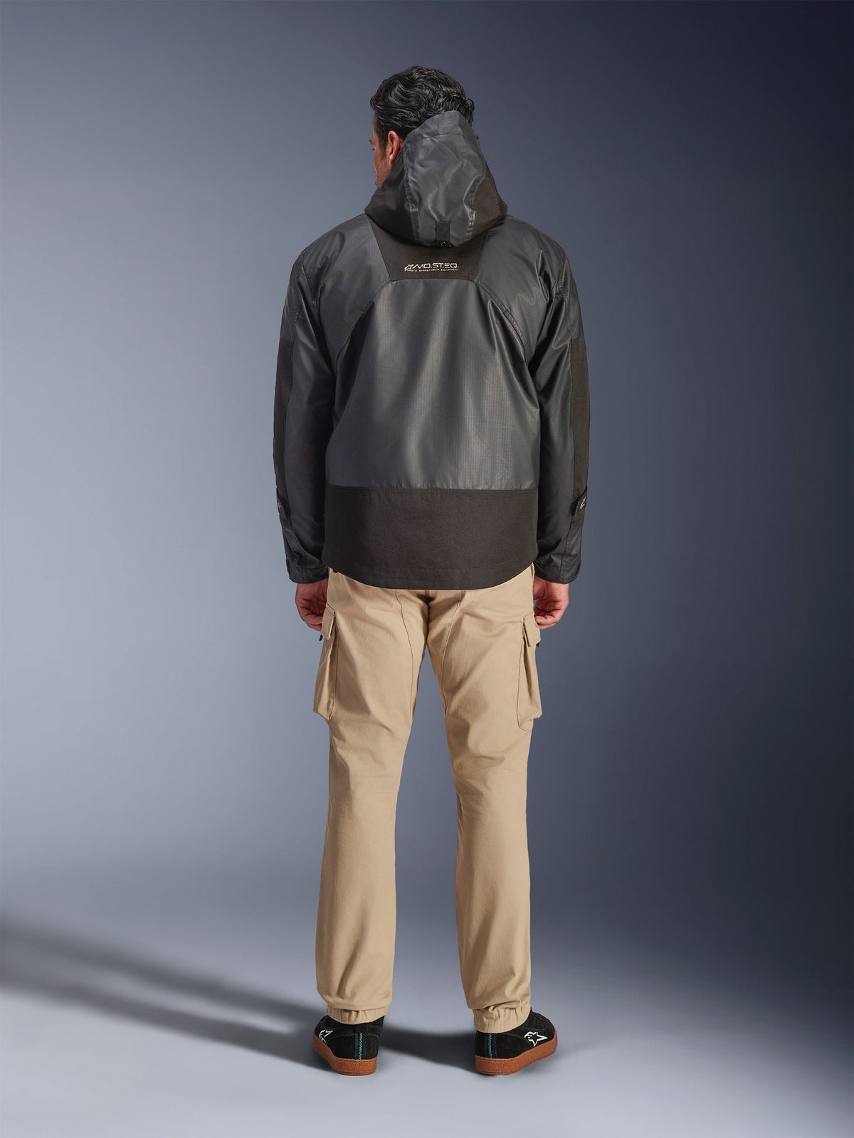 Alpinestars Mospher Waterproof Jacket, czarna parka motocyklowa widziana z tyłu, z logo MO.ST.EQ na górze pleców i konstrukcją z tkaniny ripstop, zestawiona z beżowymi spodniami cargo i czarnymi sneakersami, styl miejski.