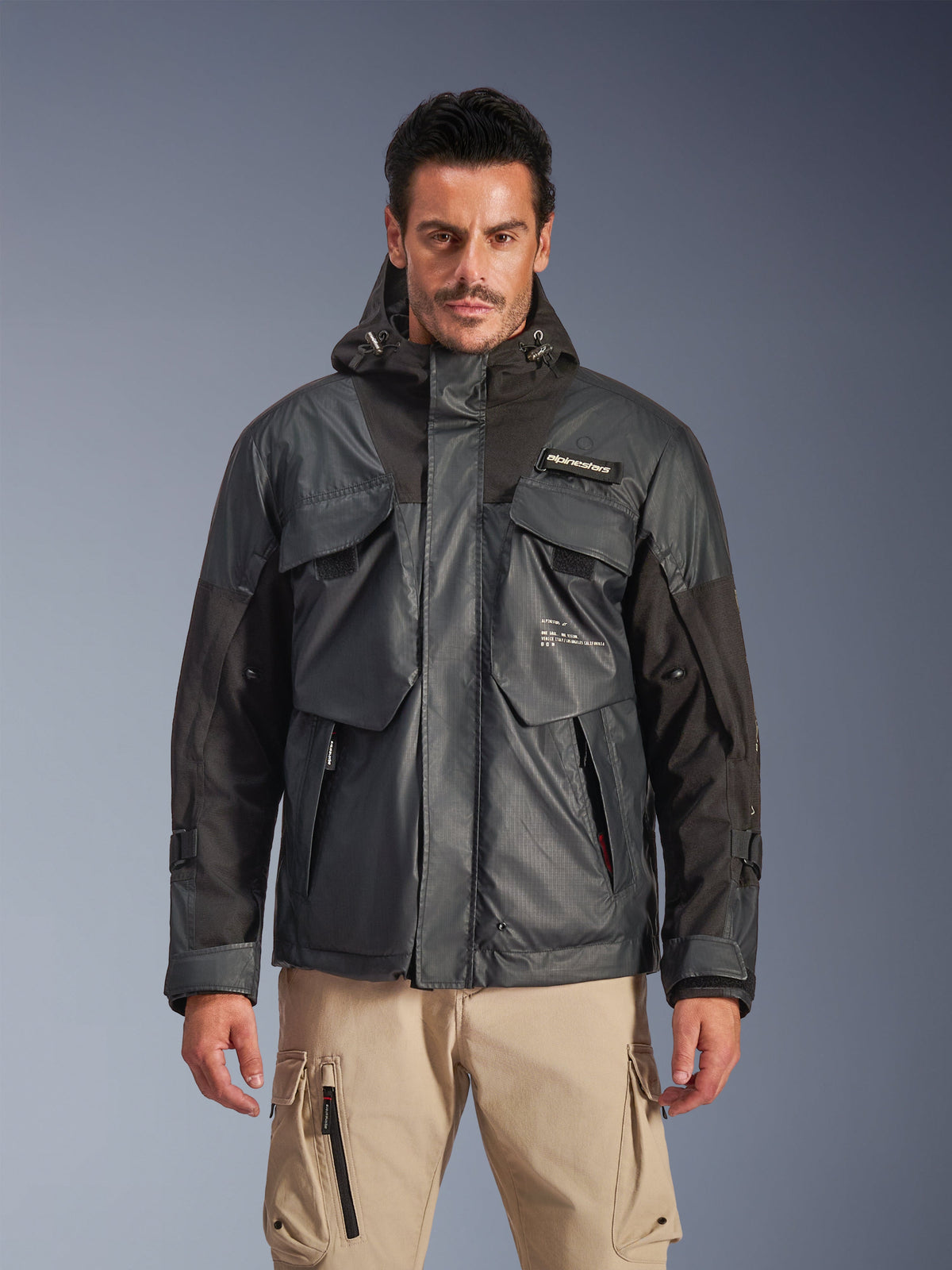 Alpinestars Mospher Waterproof Jacket, męska miejska parka motocyklowa, czarna i ciemnoszara tkanina ripstop, fason z kapturem i wieloma dużymi kieszeniami użytkowymi, prezentowana przez modela ze spodniami cargo khaki dla uzyskania taktycznego wyglądu