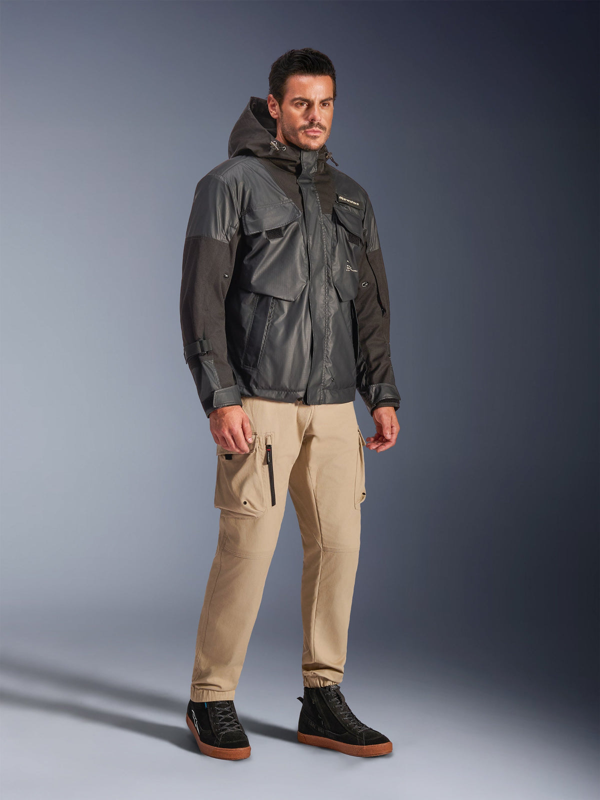 Męski model w kurtce Alpinestars Mospher Waterproof Jacket w kolorze czarnym, wodoodporna parka inspirowana stylem utility z wieloma kieszeniami i kapturem, zestawiona z beżowymi spodniami cargo i czarnymi wysokimi sneakersami, miejska odzież motocyklowa