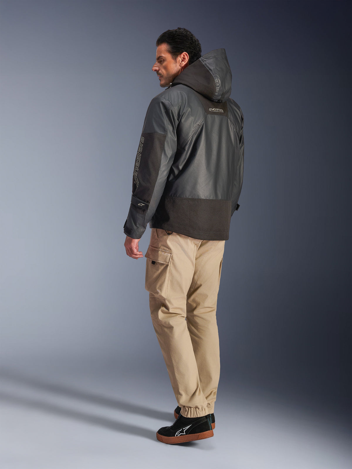 Alpinestars Mospher Waterproof Jacket w kolorze czarnym, mężczyzna widoczny z tyłu i z boku, beżowe spodnie cargo i czarne sneakersy, fason parki z kapturem, tkanina ripstop, logo Alpinestars na rękawie, nowoczesny miejski styl motocyklowy