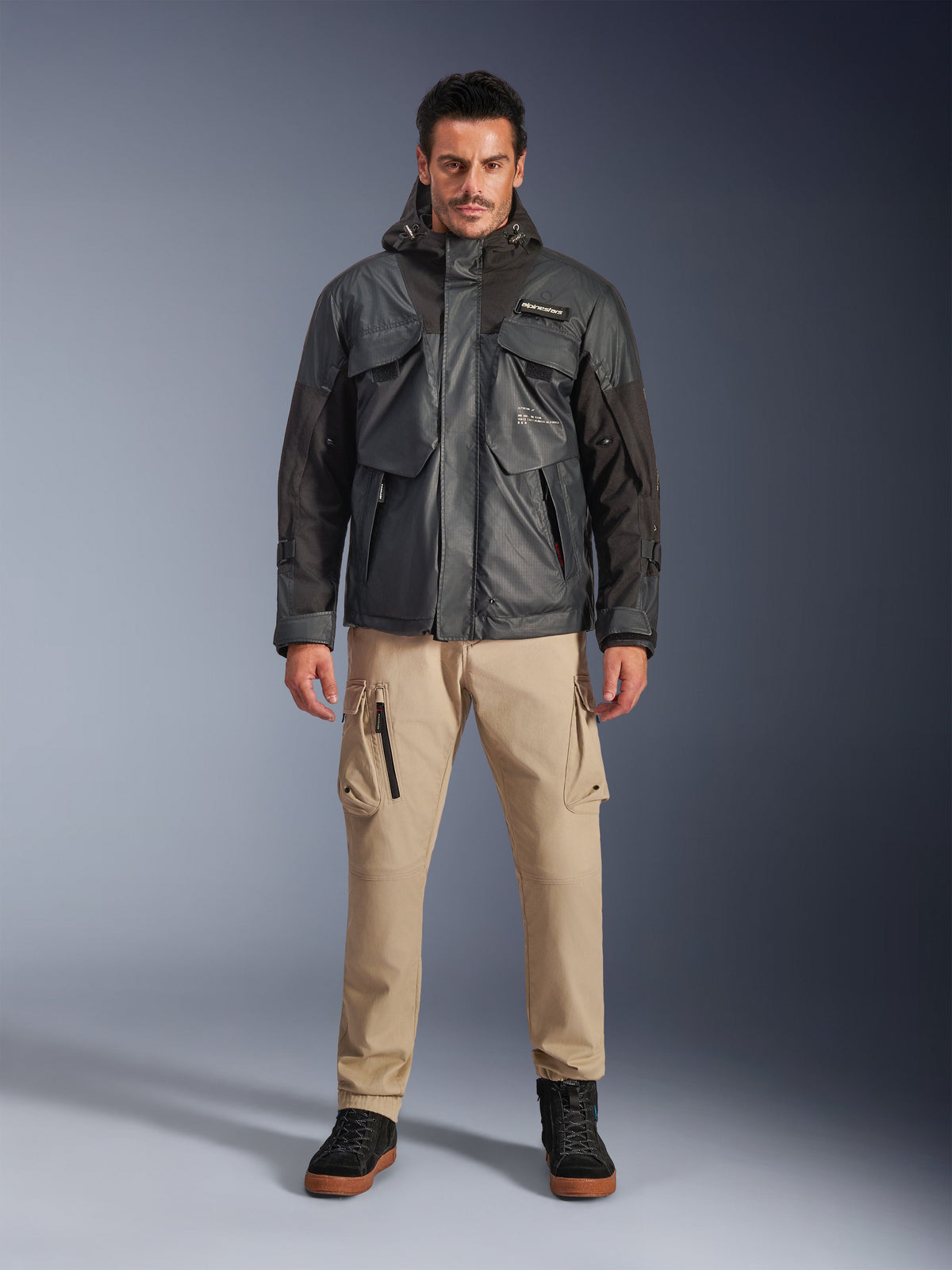 Męski model w kurtce Alpinestars Mospher Waterproof w kolorze czarnym, nowoczesna miejska odzież motocyklowa, taktyczny design użytkowy z wieloma funkcjonalnymi kieszeniami i kapturem, beżowe spodnie cargo i czarne buty, portret studyjny całej sylwetki.