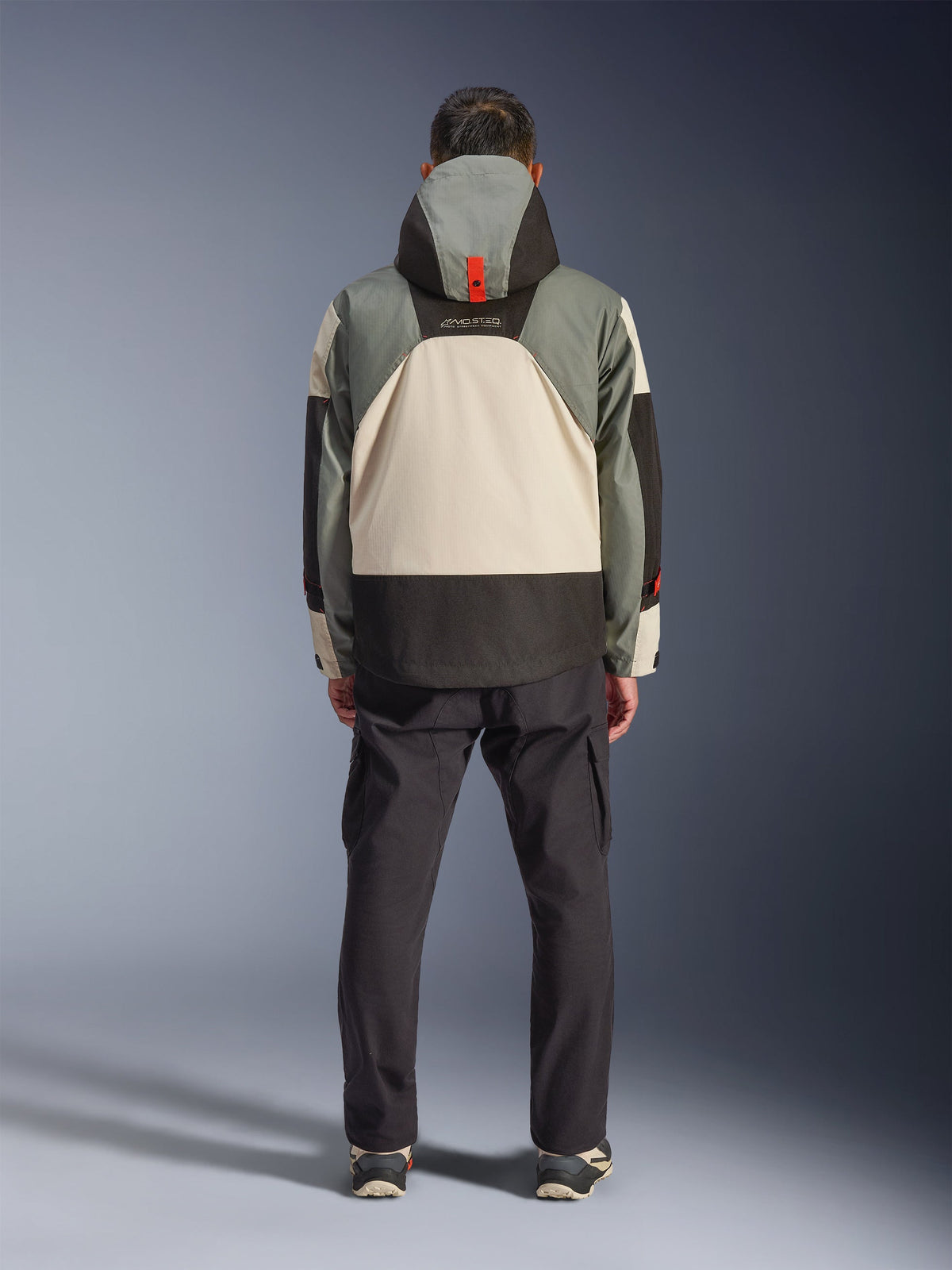 Alpinestars Mospher Waterproof Jacket, widok z tyłu motocyklisty, parka inspirowana stylem taktycznym w kolorze Fur Gray Black Ice, szare, czarne i kremowe bloki kolorów, kaptur z pomarańczowym detalem, czarne spodnie cargo