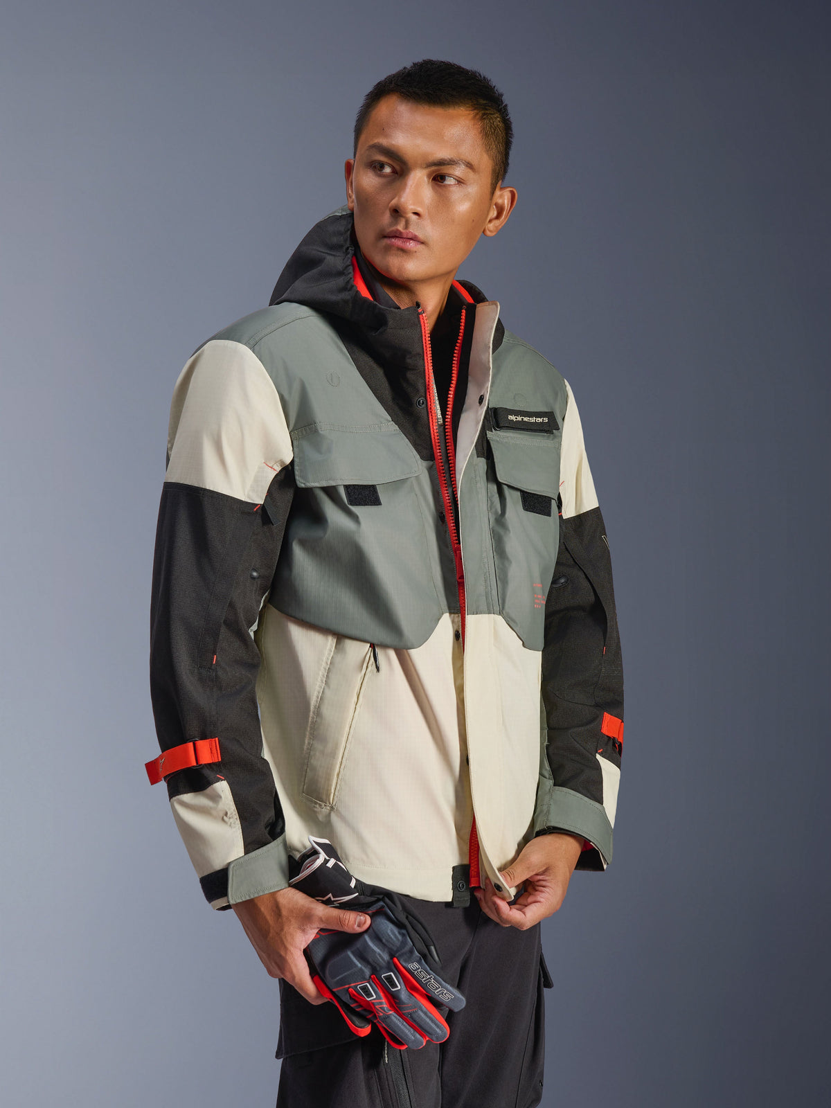 Alpinestars Mospher Waterproof Jacket w kolorze Fur Gray Black Ice, wielopanelowa tekstylna parka z szarymi, czarnymi i kremowymi sekcjami, z jaskrawoczerwonymi akcentami na suwakach i paskach, taktyczny design z kapturem i kieszeniami użytkowymi, noszona przez modela trzymającego rękawice motocyklowe.