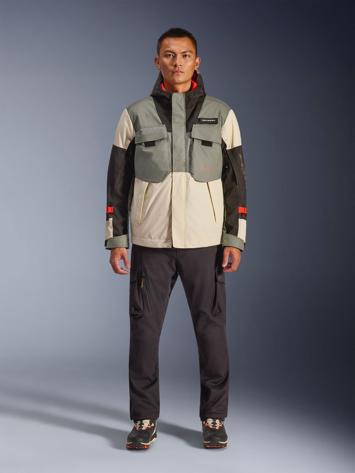 Alpinestars Mospher Waterproof Jacket w kolorze Fur Gray Black Ice, motocyklowa parka w stylu taktycznym noszona przez męskiego modela z ciemnymi spodniami cargo, konstrukcja z wieloma kieszeniami i panelami w blokach kolorystycznych szarym, kremowym i czarnym, miejska odzież motocyklowa