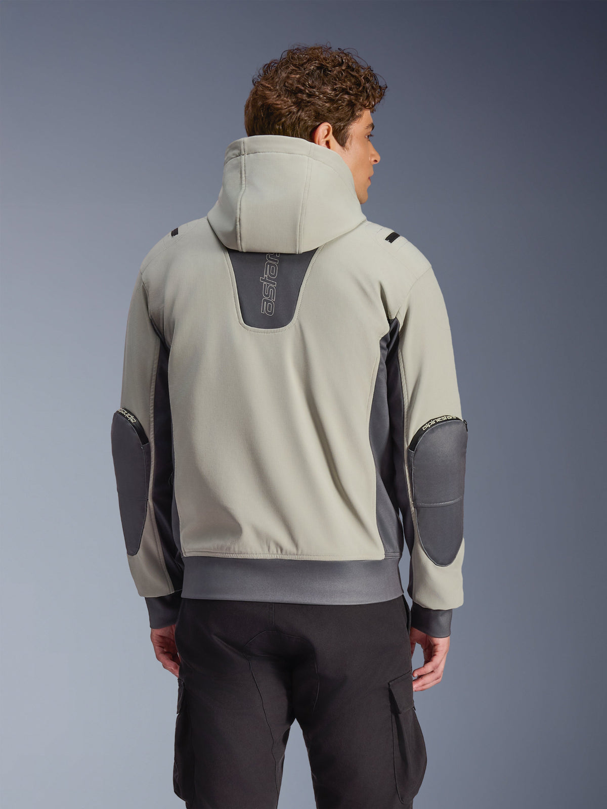 Kurtka Alpinestars Moblast Waterproof w kolorze Iron Gray i Dark Sage, widok z tyłu na modelu, miejska wodoodporna kurtka motocyklowa z kapturem, ciemnoszare elastyczne panele i wzmocnione łokcie, zestawiona z czarnymi spodniami cargo