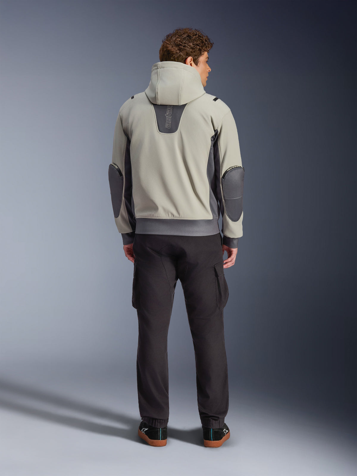Alpinestars Moblast Waterproof Jacket, kolor Iron Gray Dark Sage, widok z tyłu na motocyklistę w kurtce motocyklowej z kapturem, jasnoszara i ciemnoszałwiowa kolorystyka, pionowe logo „astars” w górnej części pleców, ciemnoszare łaty na łokciach, stylizacja z ciemnymi spodniami cargo dla nowoczesnego miejskiego wyglądu.