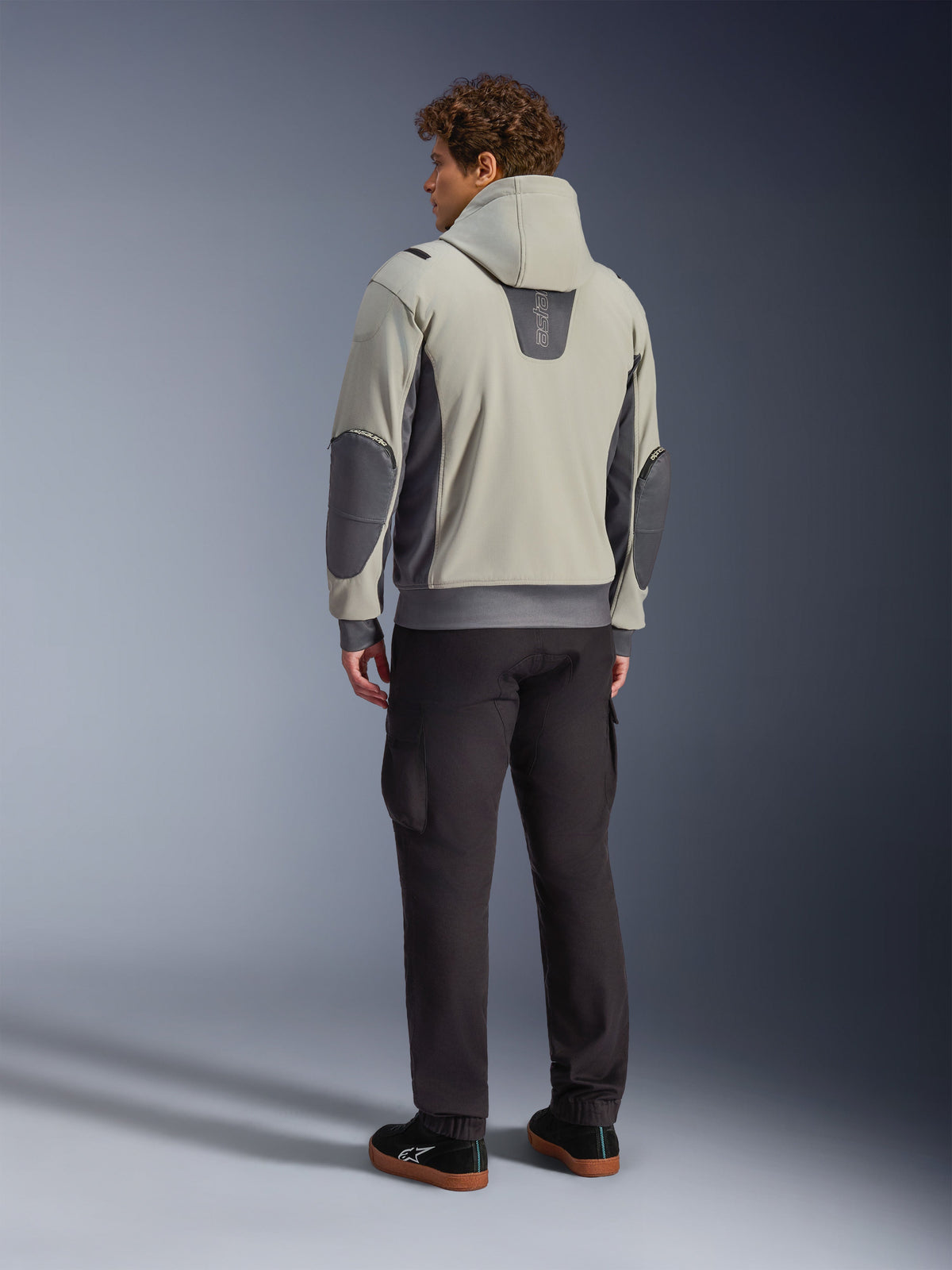 Męski model widoczny od tyłu w kurtce Alpinestars Moblast Waterproof Jacket w kolorze Iron Gray Dark Sage, miejska kurtka motocyklowa z kapturem, ciemnoszarymi panelami bocznymi i wzmocnionymi łatami na łokciach, w zestawieniu z ciemnymi spodniami motocyklowymi i czarnymi tenisówkami, nowoczesna minimalistyczna estetyka do miejskich dojazdów