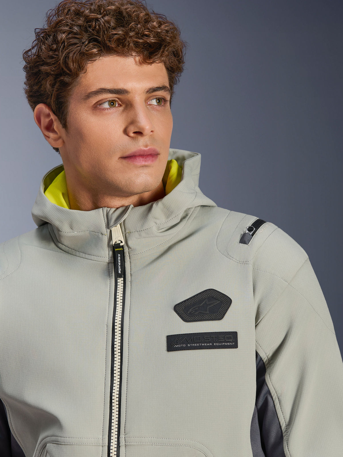 Kurtka Alpinestars Moblast Waterproof, Iron Gray Dark Sage, zbliżenie na modela w kurtce, czarne logo Alpinestars i naszywki MO.ST.EQ. na klatce piersiowej, jaskrawożółta podszewka kaptura, nowoczesny techniczny design.