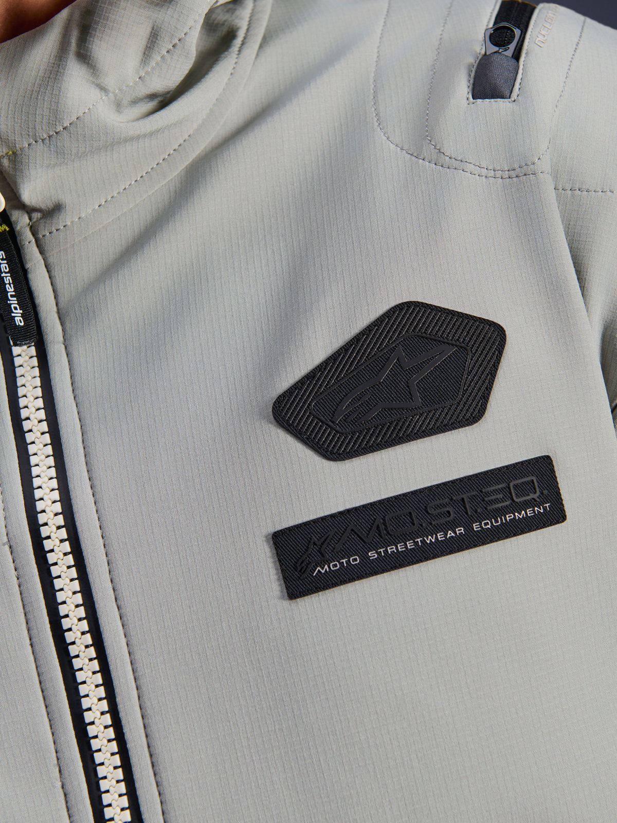 Kurtka Alpinestars Moblast Waterproof, Iron Gray, zbliżenie na naszywki na klatce piersiowej z logo Alpinestars i brandingiem MO.ST.EQ. Moto Streetwear Equipment, techniczna tkanina ripstop, solidny zamek błyskawiczny oraz detal kieszeni na ramieniu.