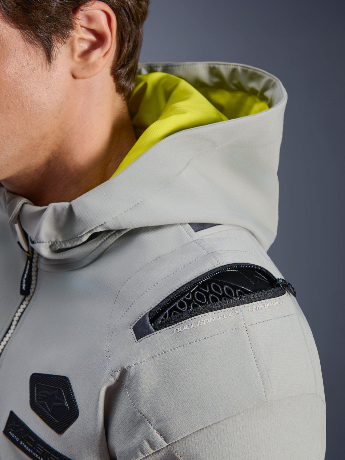 Kurtka Alpinestars Moblast WP, kolor Iron Gray, zbliżenie na ramię z otwartą komorą na ochraniacz Nucleon Flex, neonowożółte wnętrze kaptura, techniczna miejska odzież motocyklowa z funkcjonalnymi zamkami