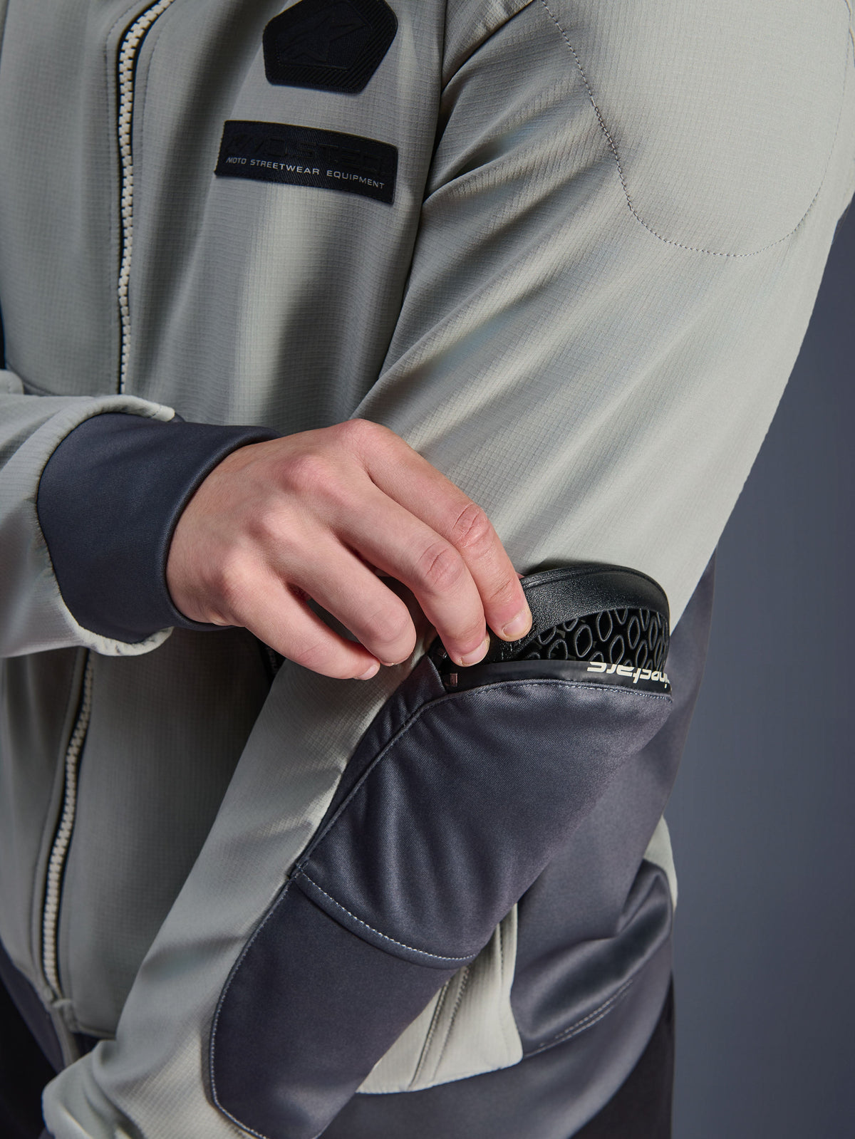 Wodoodporna kurtka Alpinestars Moblast, kolor Iron Gray i Dark Sage, zbliżenie pokazujące wyjmowanie ochraniacza Nucleon Flex Plus z zewnętrznej kieszeni na łokciu zapinanej na zamek, techniczna miejska odzież motocyklowa z brandingiem MO.ST.EQ.