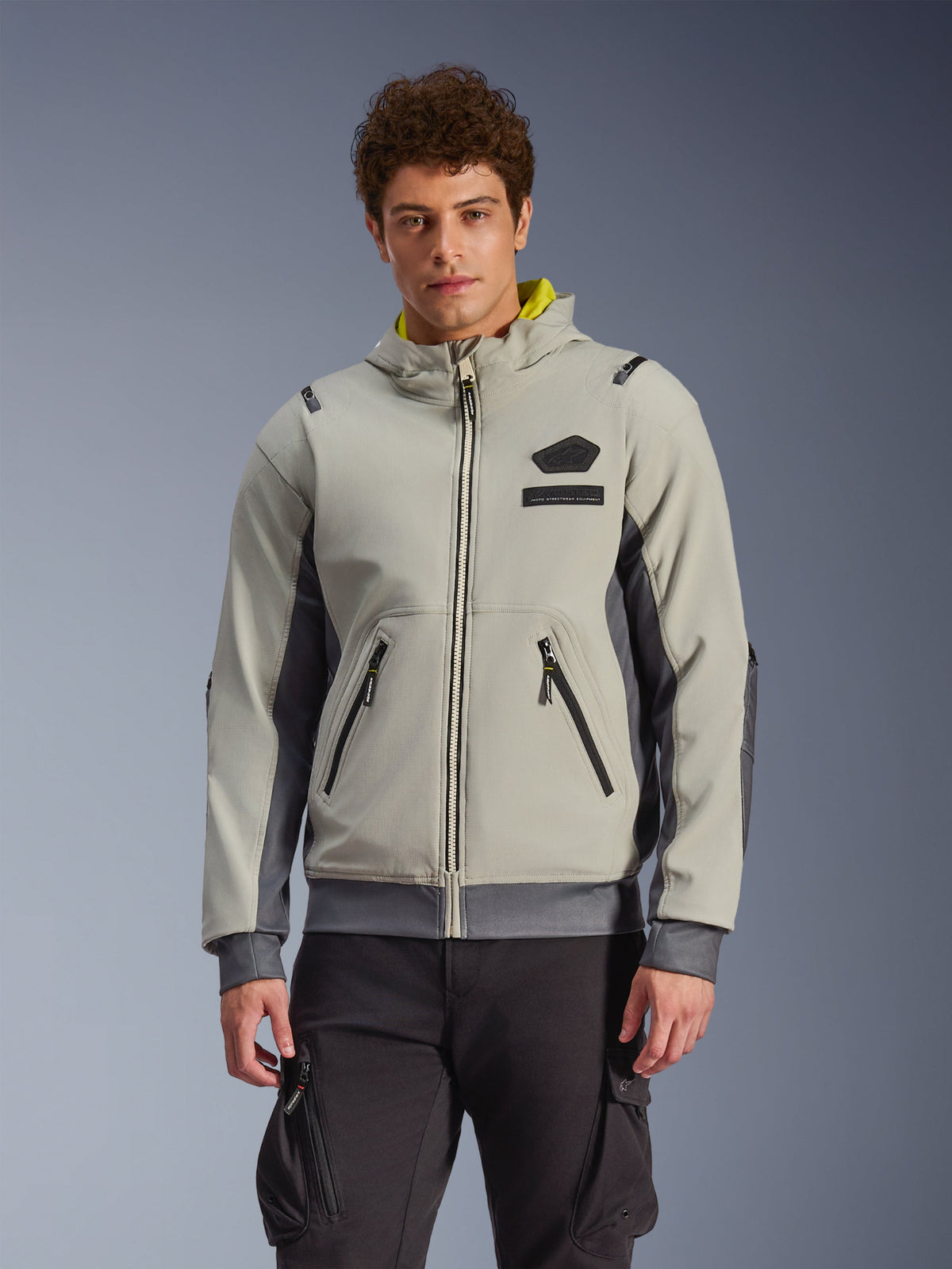 Alpinestars Moblast Waterproof Jacket, jasnoszara i ciemnoszara miejska bluza motocyklowa z żółtą podszewką kaptura, przednimi kieszeniami i czarnymi zamkami, noszona z czarnymi spodniami bojówkami, nowoczesny sportowy design