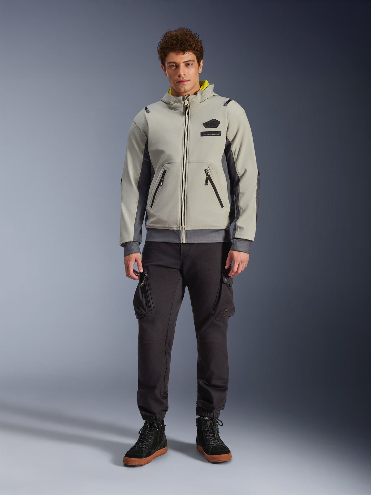 Męski model w kurtce Alpinestars Moblast Waterproof Jacket w kolorze Iron Gray i Dark Sage, motocyklowa kurtka z kapturem o nowoczesnym technicznym designie, zestawiona z ciemnymi spodniami motocyklowymi cargo i czarnymi butami, miejska odzież motorsportowa