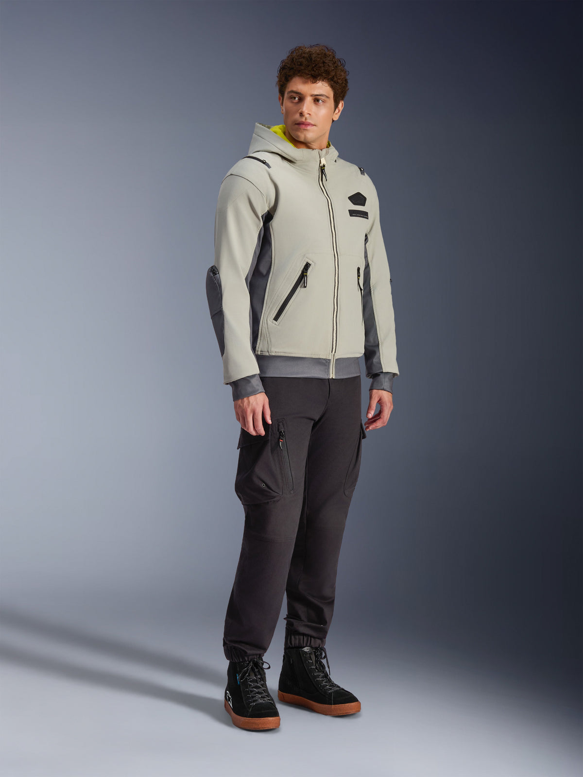 Alpinestars Moblast Waterproof Jacket, Iron Gray i Dark Sage, miejska kurtka motocyklowa o fasonie bluzy z kapturem, prezentowana przez modela w ciemnych spodniach i czarnych butach, minimalistyczna estetyka cyber-tech z żółtym wnętrzem kaptura i wodoodpornymi zamkami